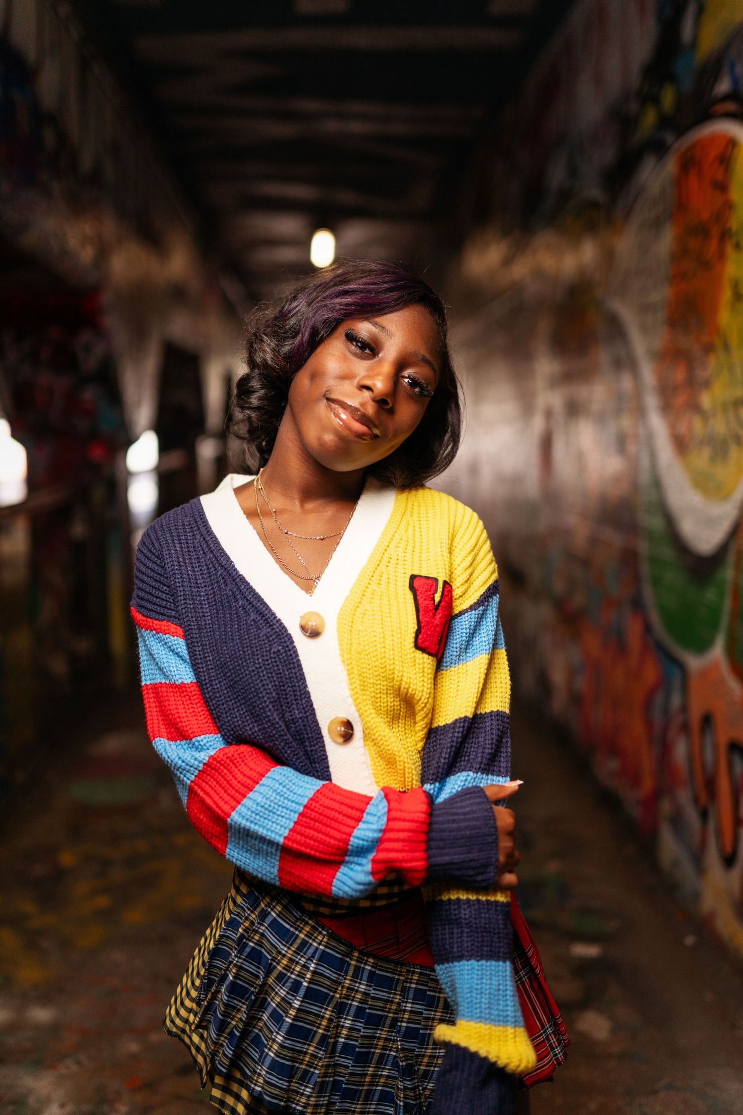 Fashion model poses in colorful patchwork sweater against urban graffiti backdrop.