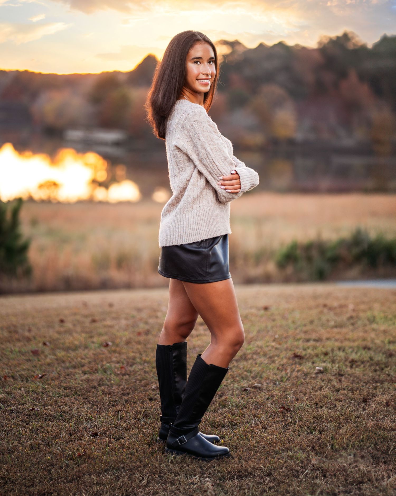 Person in cream sweater and boots poses in field during golden hour sunset.