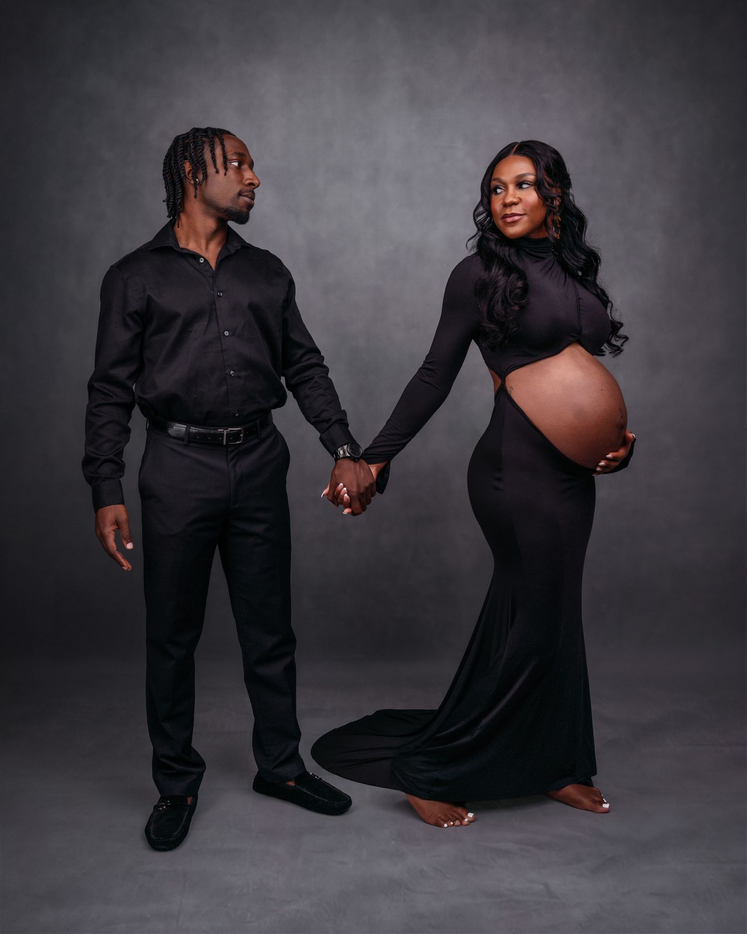 Maternity photo series with couple in all black outfits against grey backdrop.