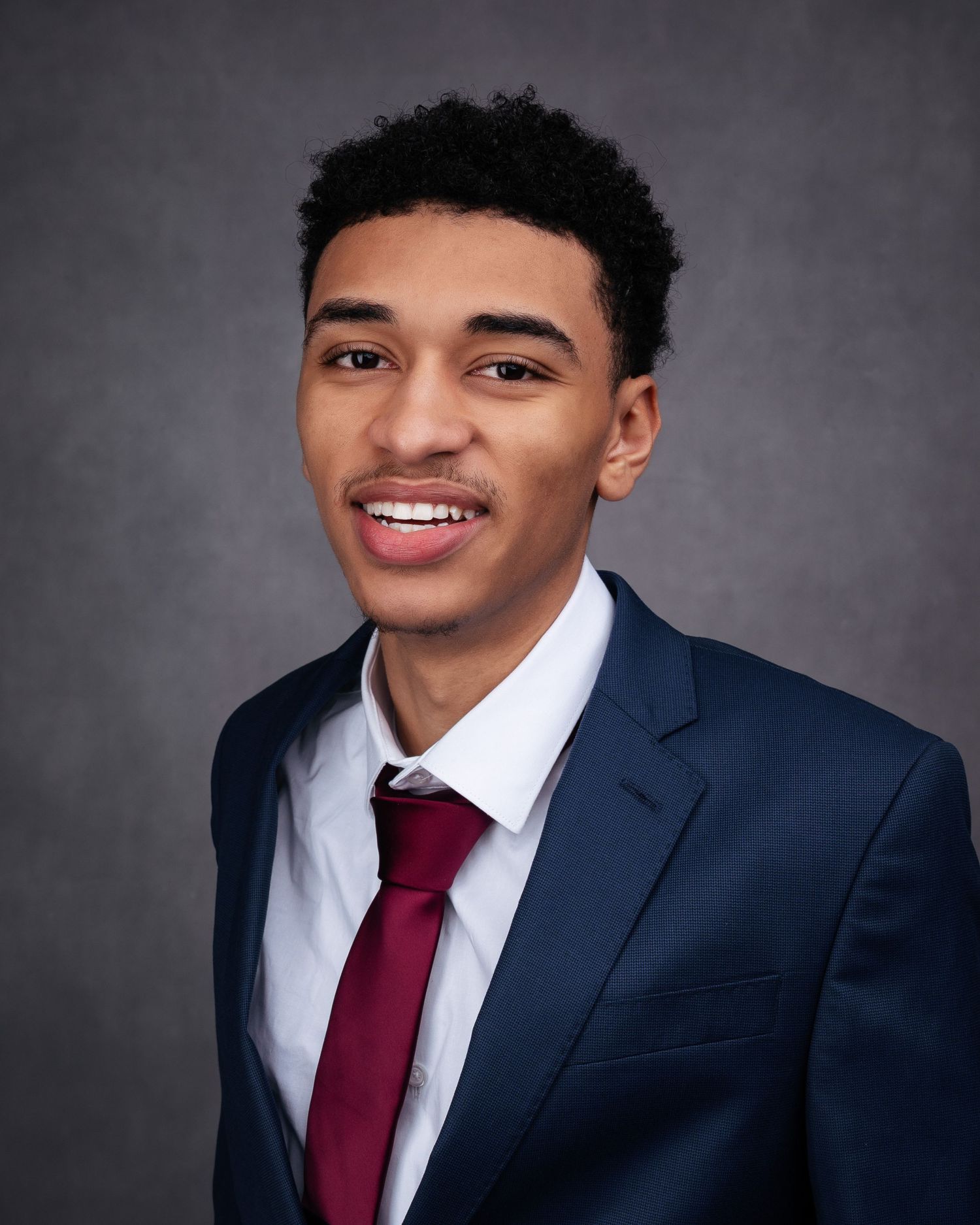 Professional headshots against grey background featuring navy suit and red tie.