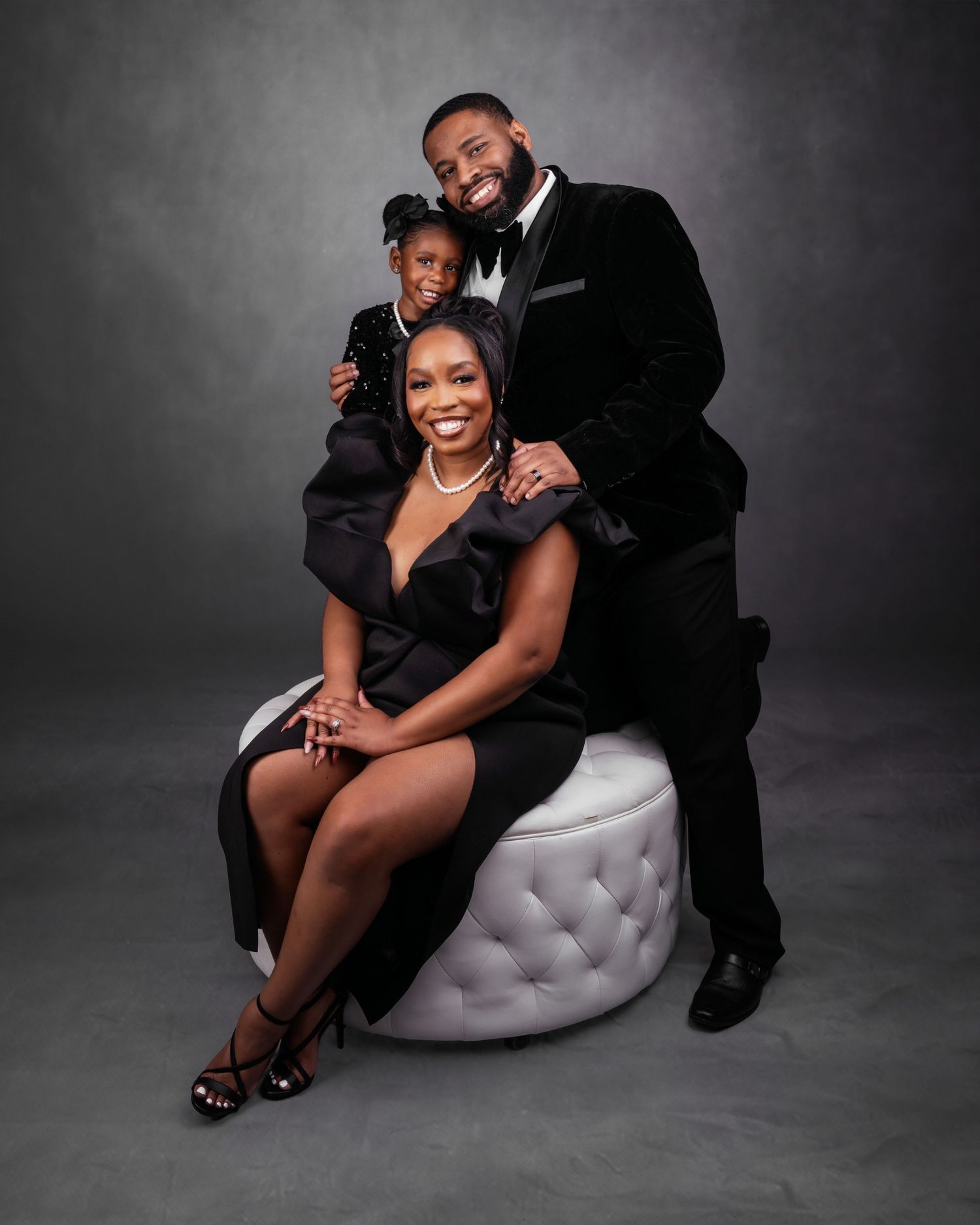 A formal studio portrait featuring elegant black attire and a white tufted ottoman.