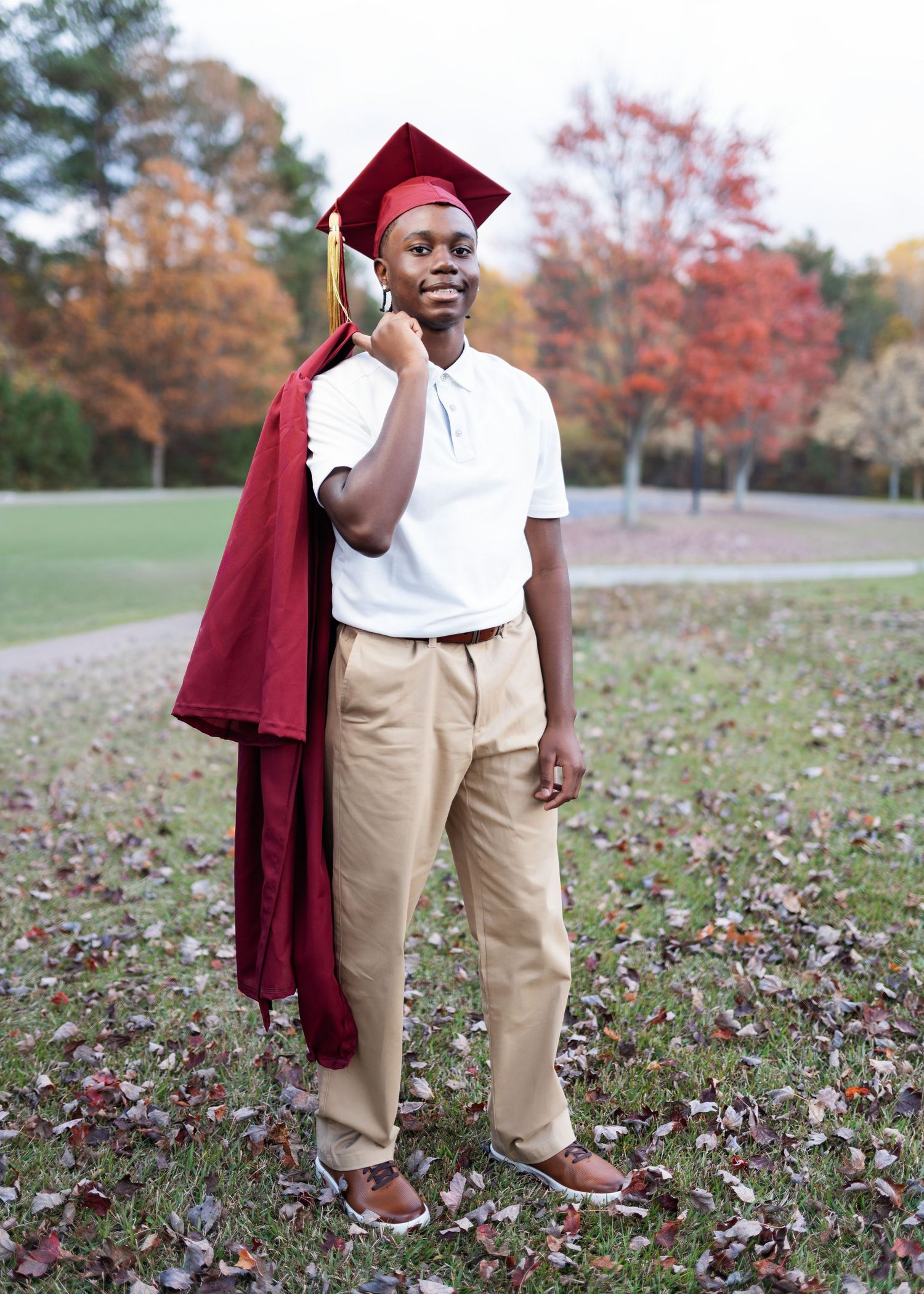 Person in graduation attire poses playfully in field with autumn leaves.
