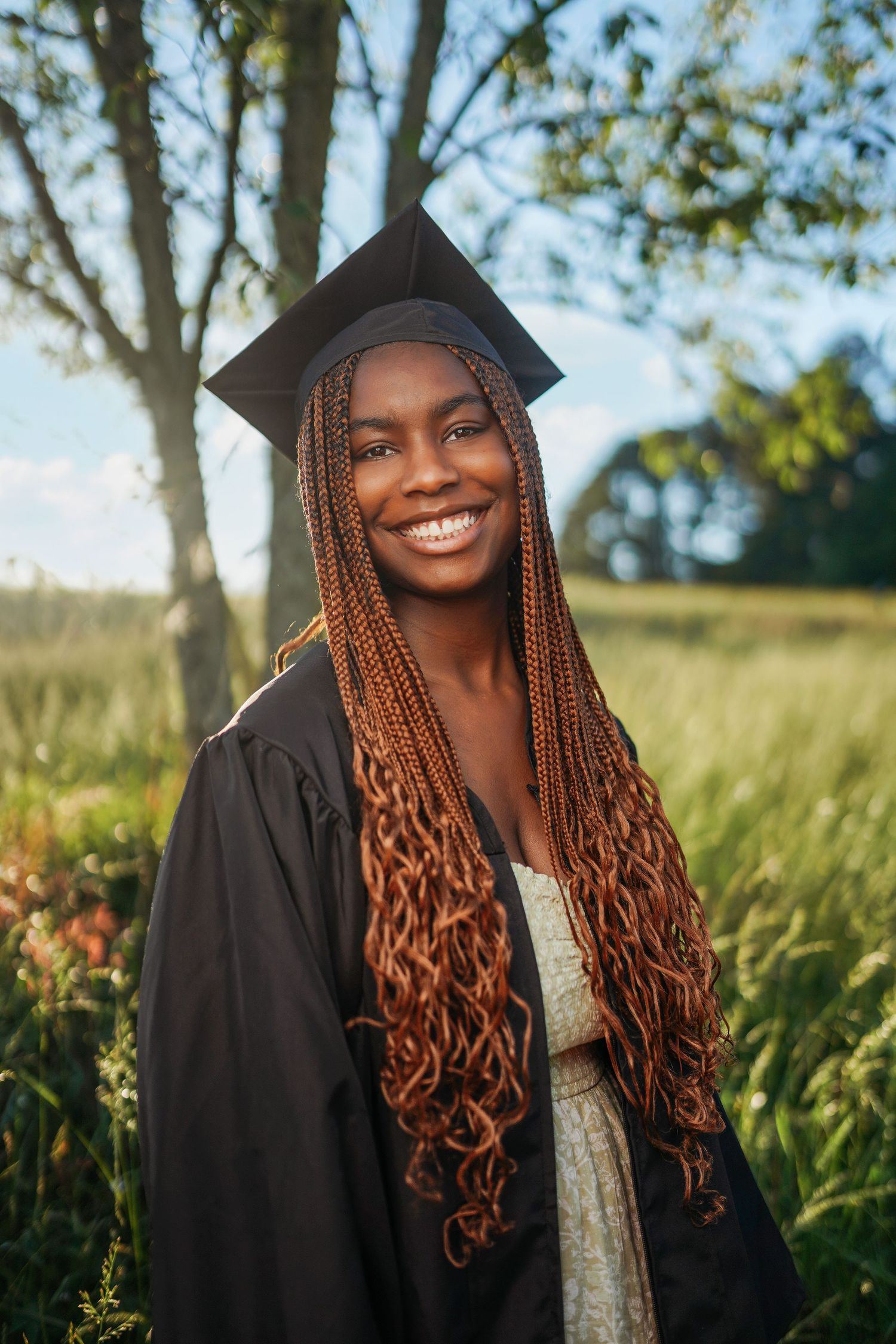 Graduate in black cap and gown with long braided hair poses outdoors in a sunny field.