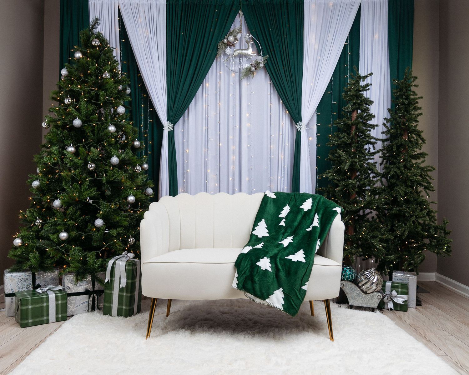 Cream boucle loveseat with green Christmas tree blanket, decorated trees, and wrapped gifts on a white rug before emerald green curtains.