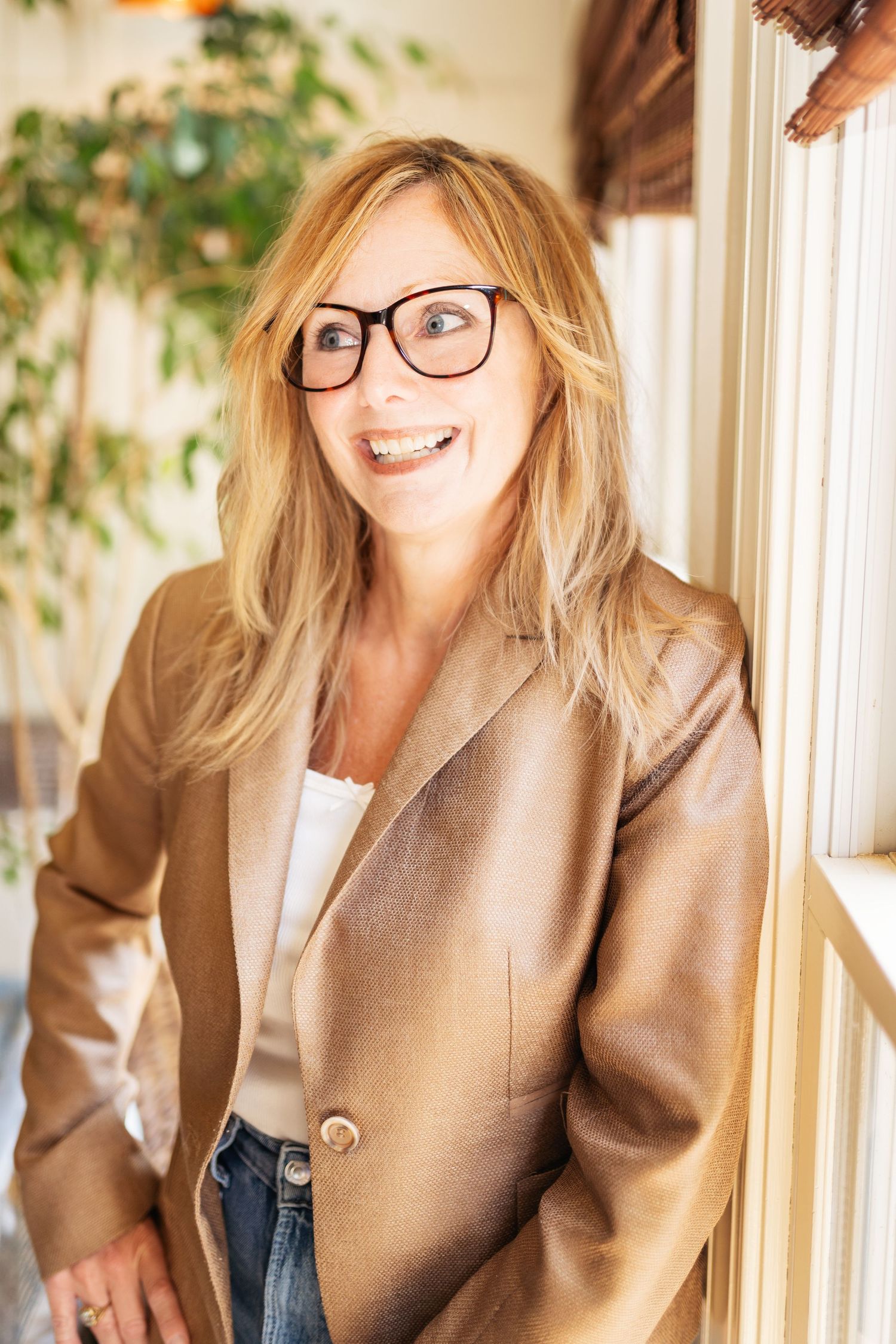Person in tan blazer posing by window in bright interior setting.