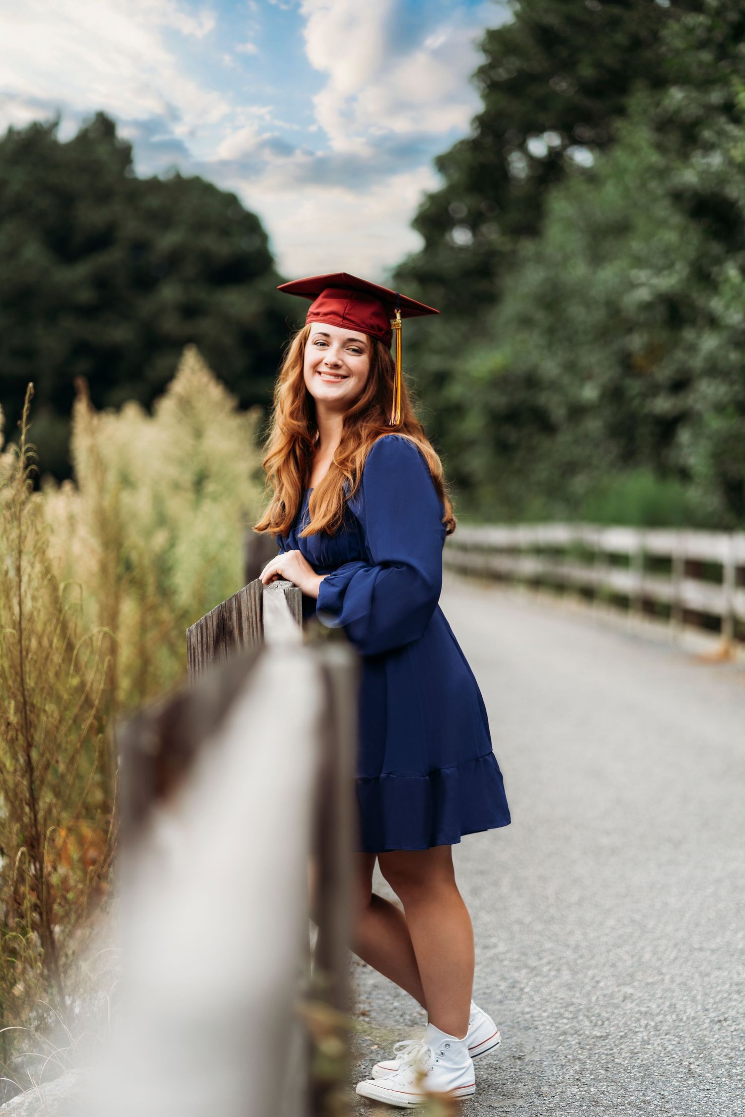 Graduate in red cap and gown poses confidently on path with fence and tall grass.