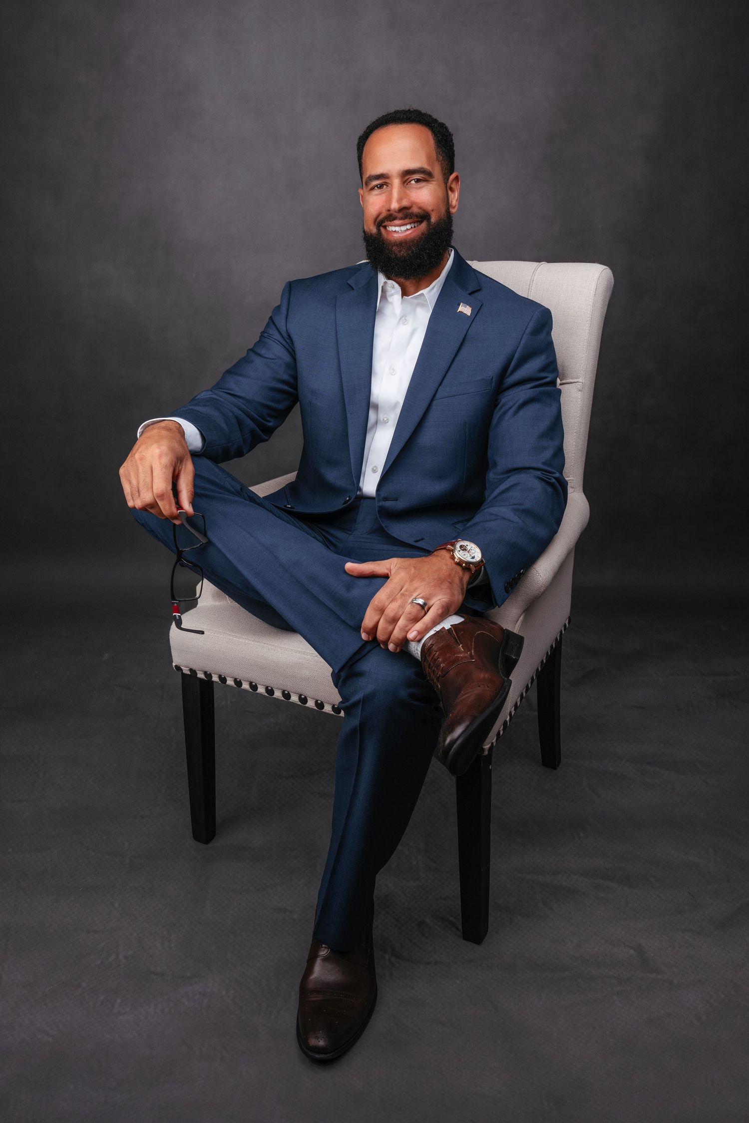 Professional portrait of a businessman in a navy suit seated confidently in a gray armchair against dark background.