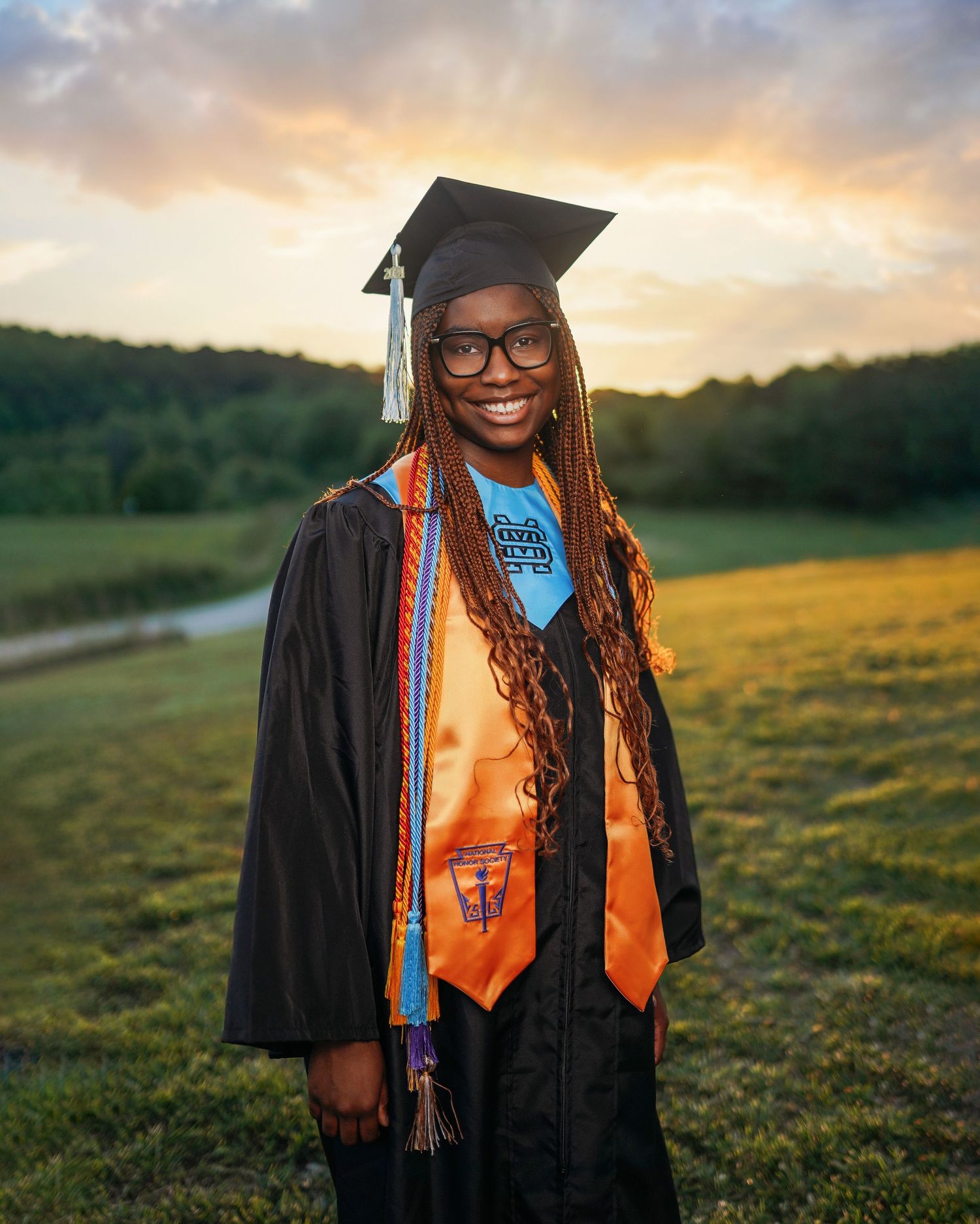 Graduate in cap and gown with orange stole smiling during sunset photo session.