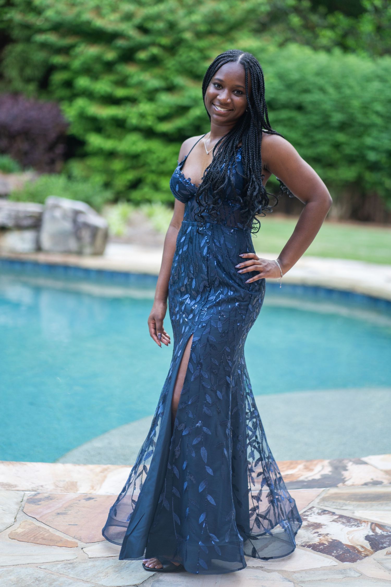 Person in navy blue formal dress posing by poolside with lush greenery in background.