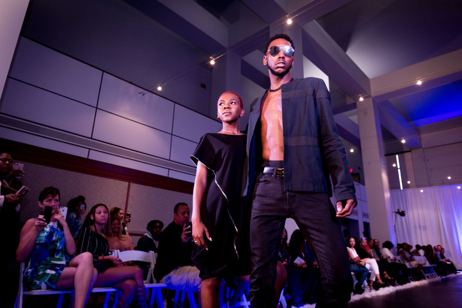 Models perform on darkened runway stage with dramatic lighting and seated audience.