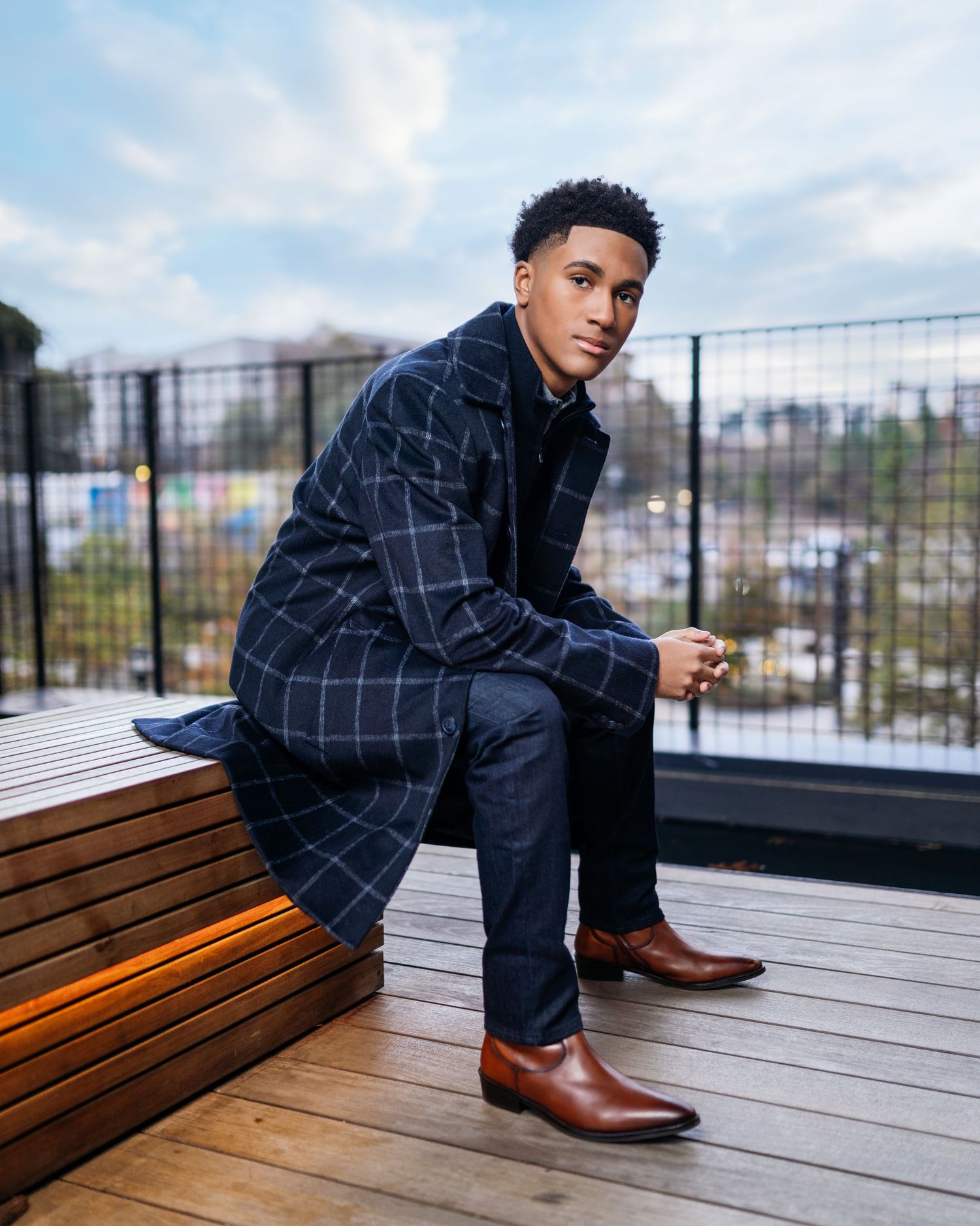 Professional poses on wooden deck wearing plaid jacket and brown boots.