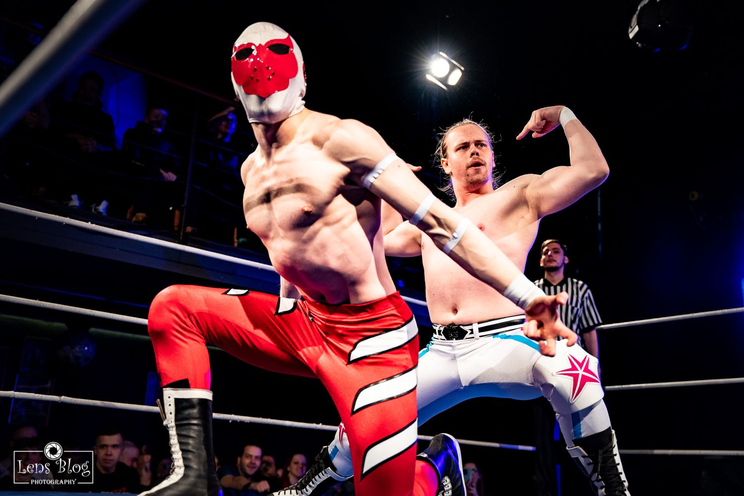 Two wrestlers engage in combat with one wearing red attire in a brightly lit wrestling ring.