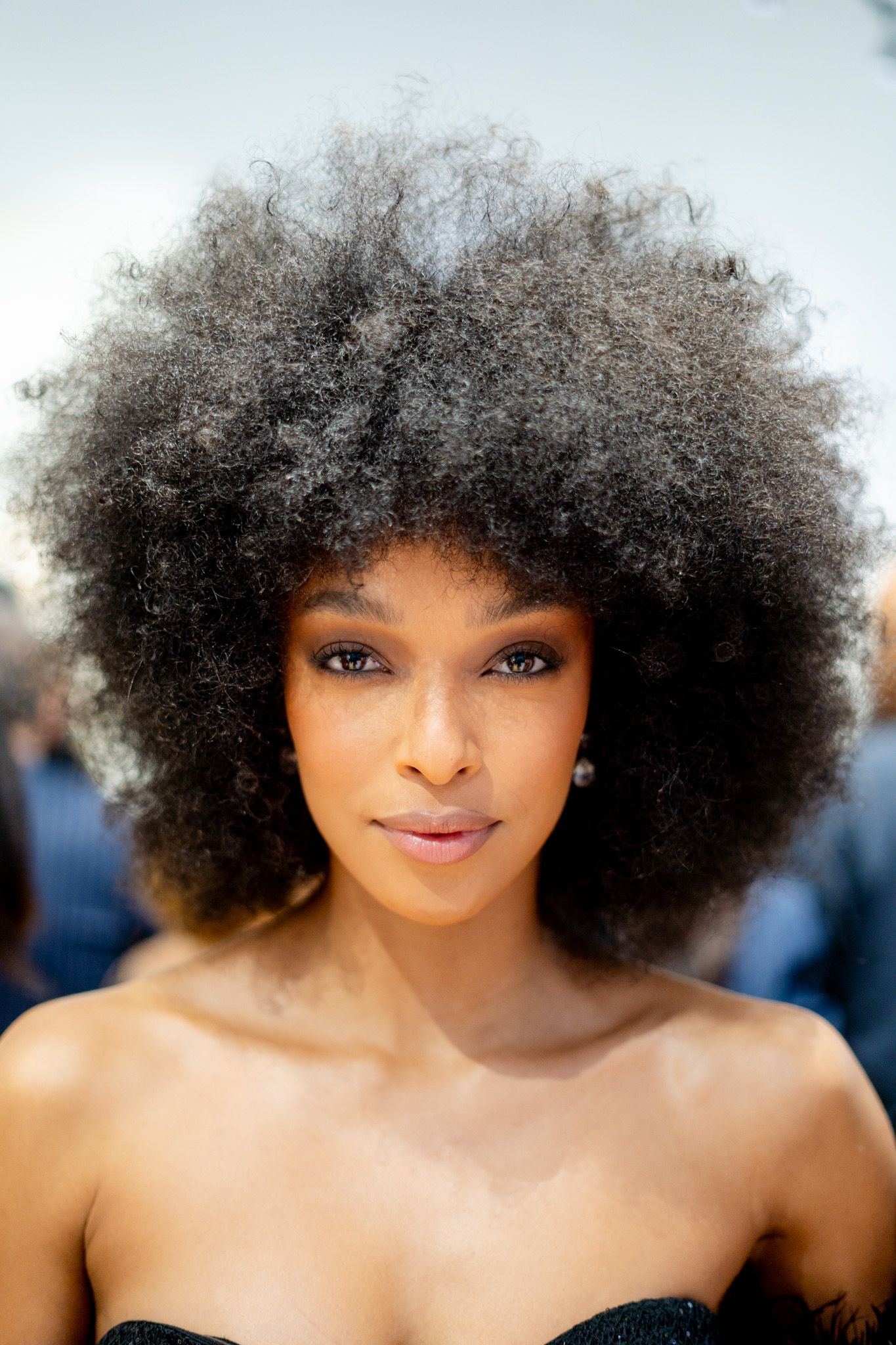 A young Black woman with a voluminous natural afro hairstyle poses confidently, wearing a strapless black top.