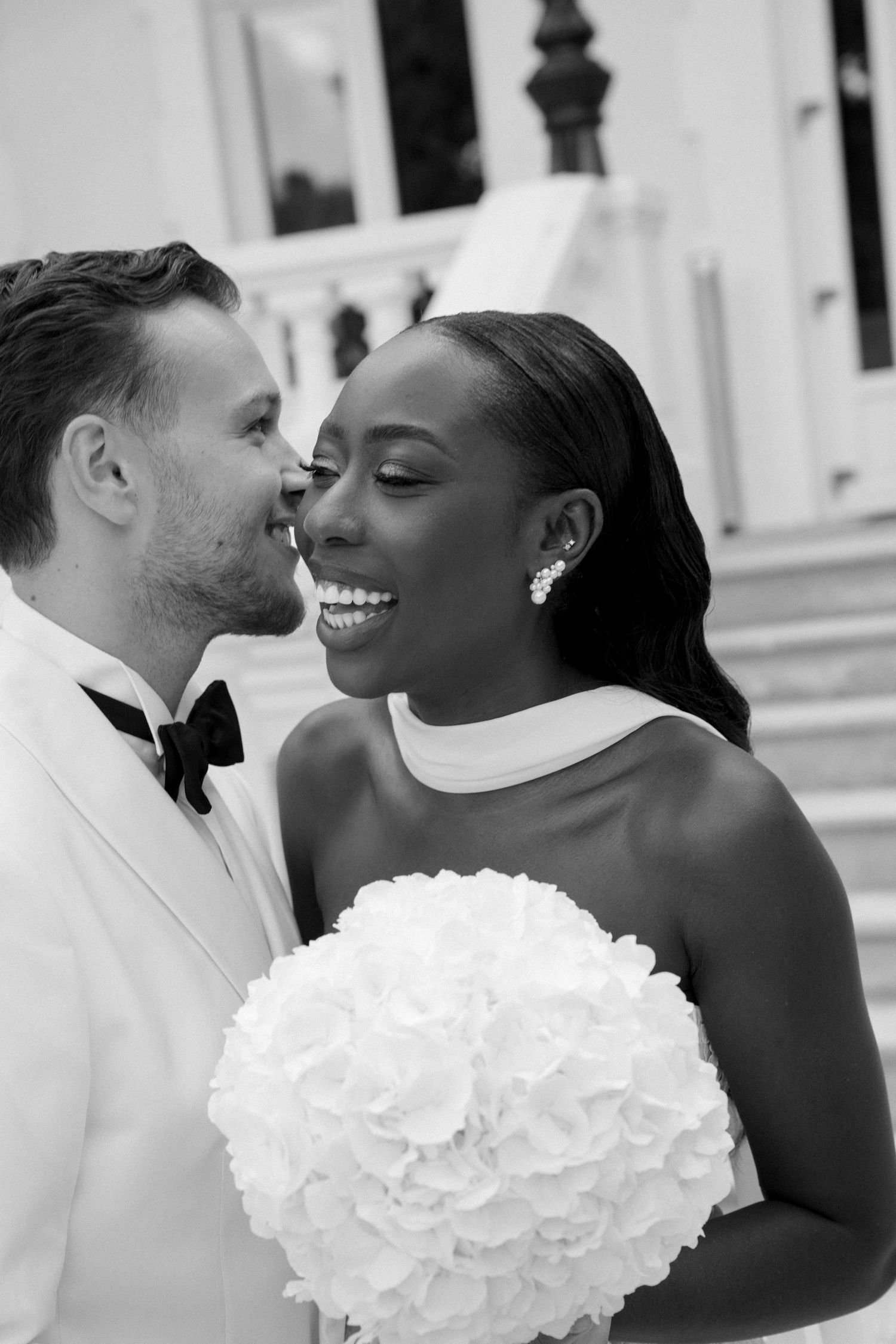 Black and white wedding photo of smiling interracial couple, bride holding white bouquet, groom in white tuxedo with bow tie.