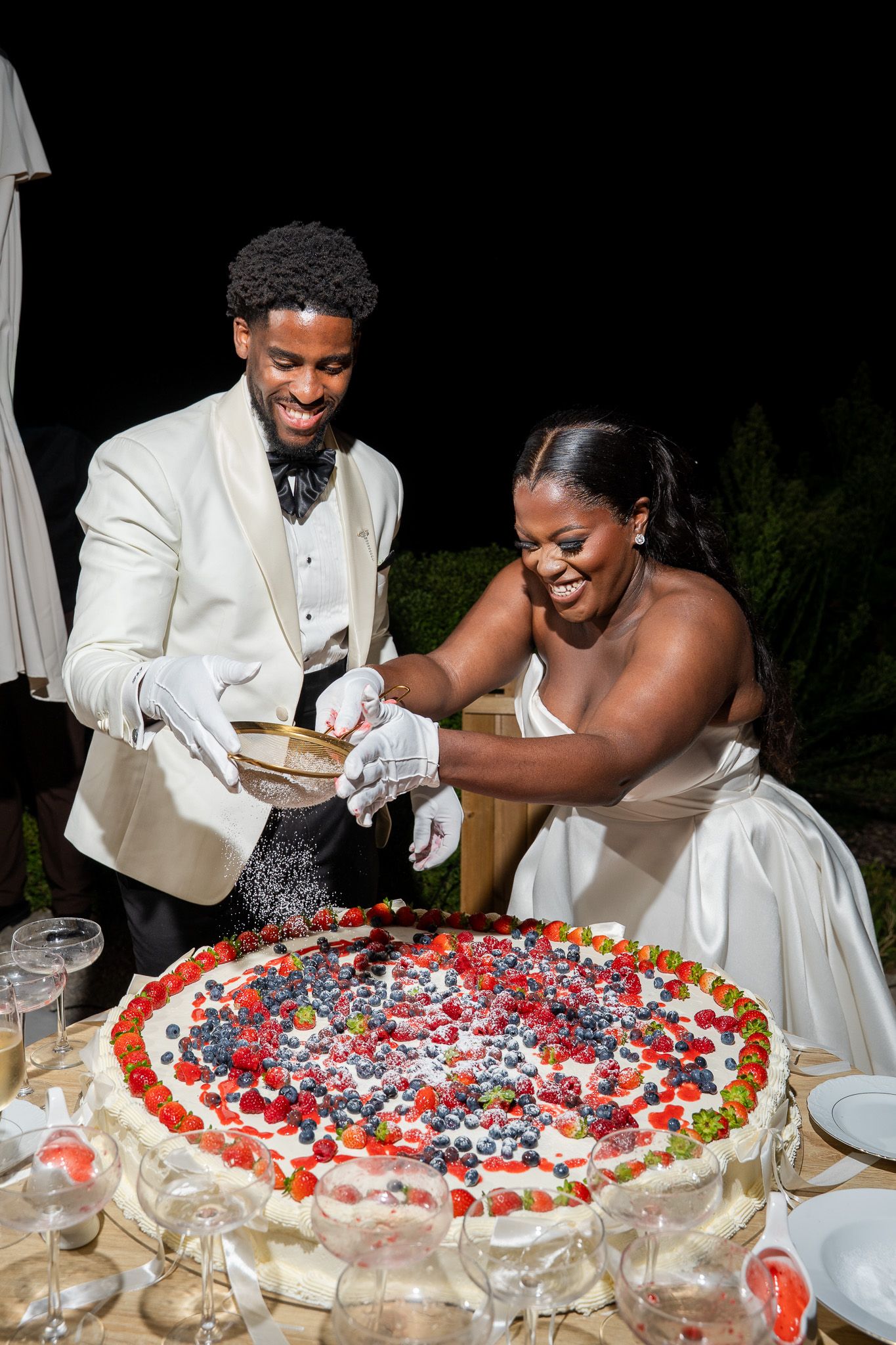 Bride and groom cutting a large fruit-topped wedding cake outdoors at night, both smiling and wearing white gloves.