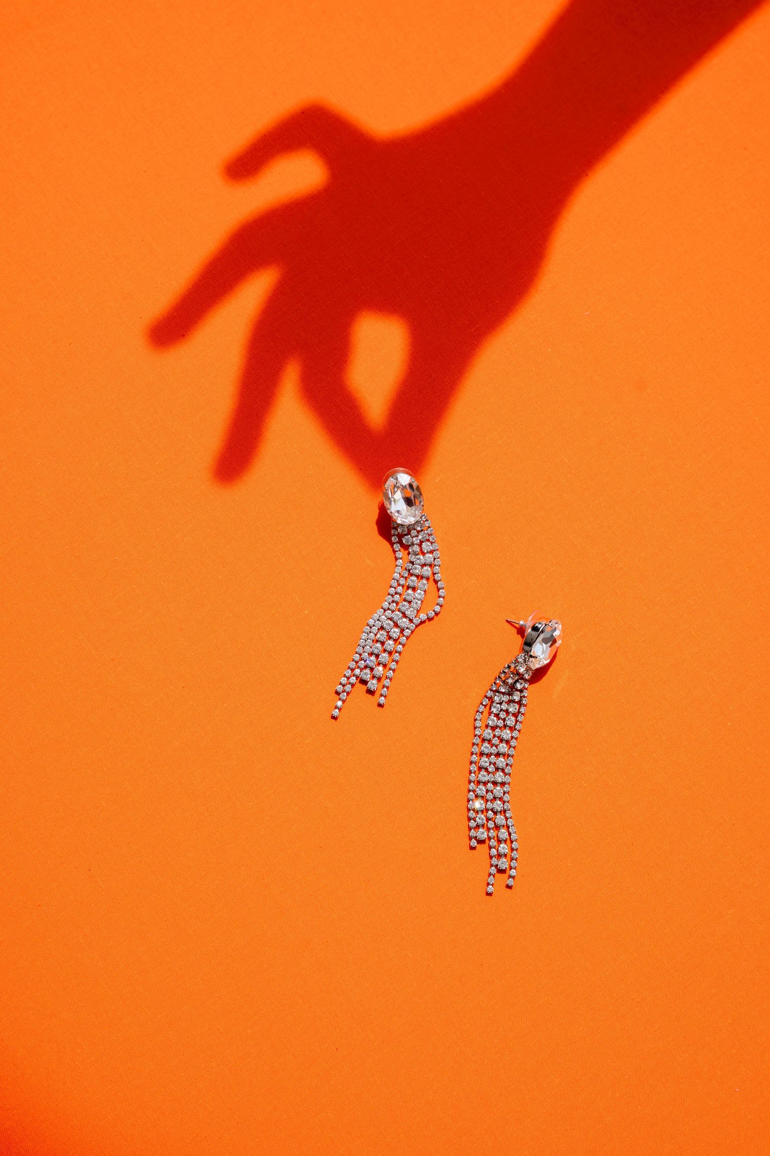 Crystal tassel drop earrings on orange background with dramatic hand shadow reaching for jewelry.
