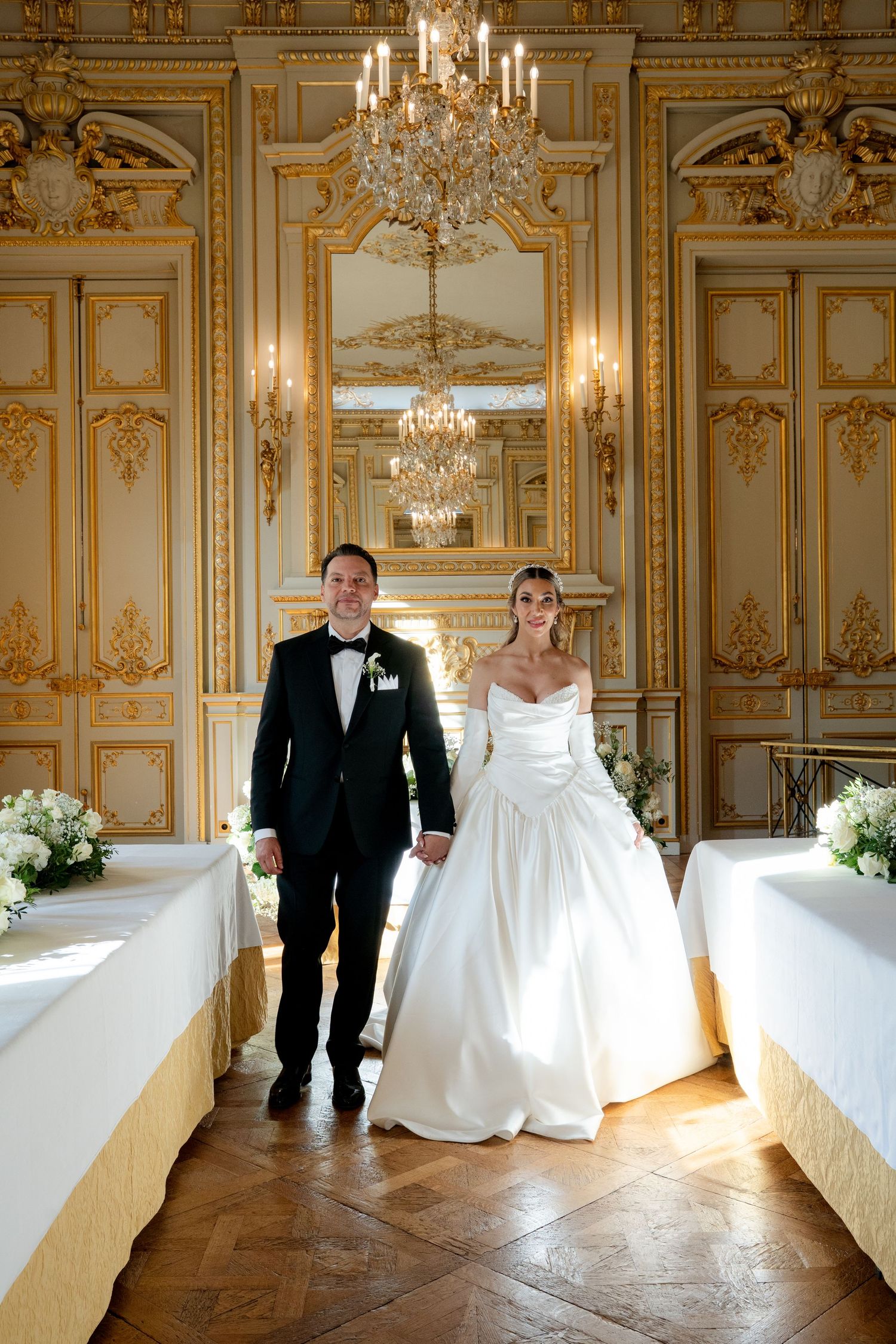 Bride and groom walk hand-in-hand in an ornate gold Parisian ballroom with crystal chandeliers on their wedding day.