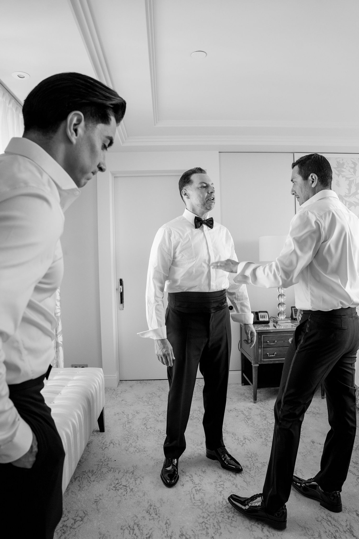 Black and white photo of three men in formal attire preparing for a wedding ceremony in an elegant room.