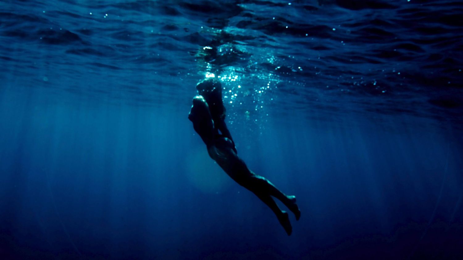 Silhouetted scuba diver swimming in deep blue ocean waters with sunlight streaming from above.