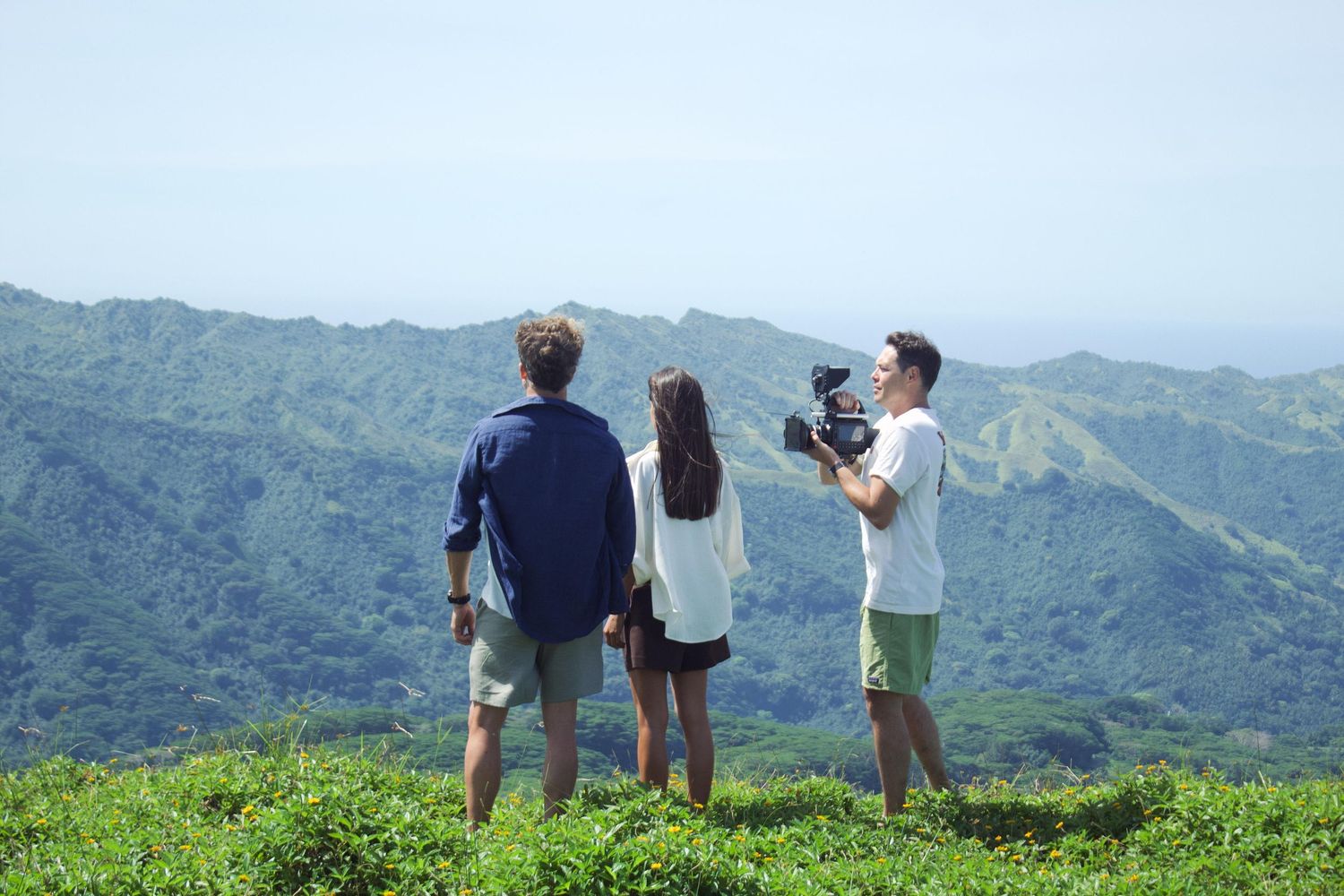 Three people filming lush green mountain landscape from a scenic overlook on a clear day.
