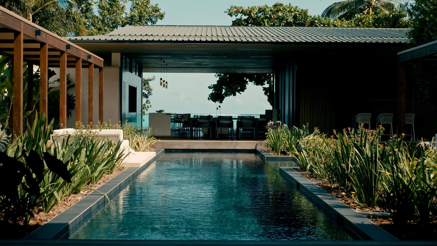 Luxury villa with a serene rectangular pool surrounded by tropical plants, open pavilion, and ocean view.