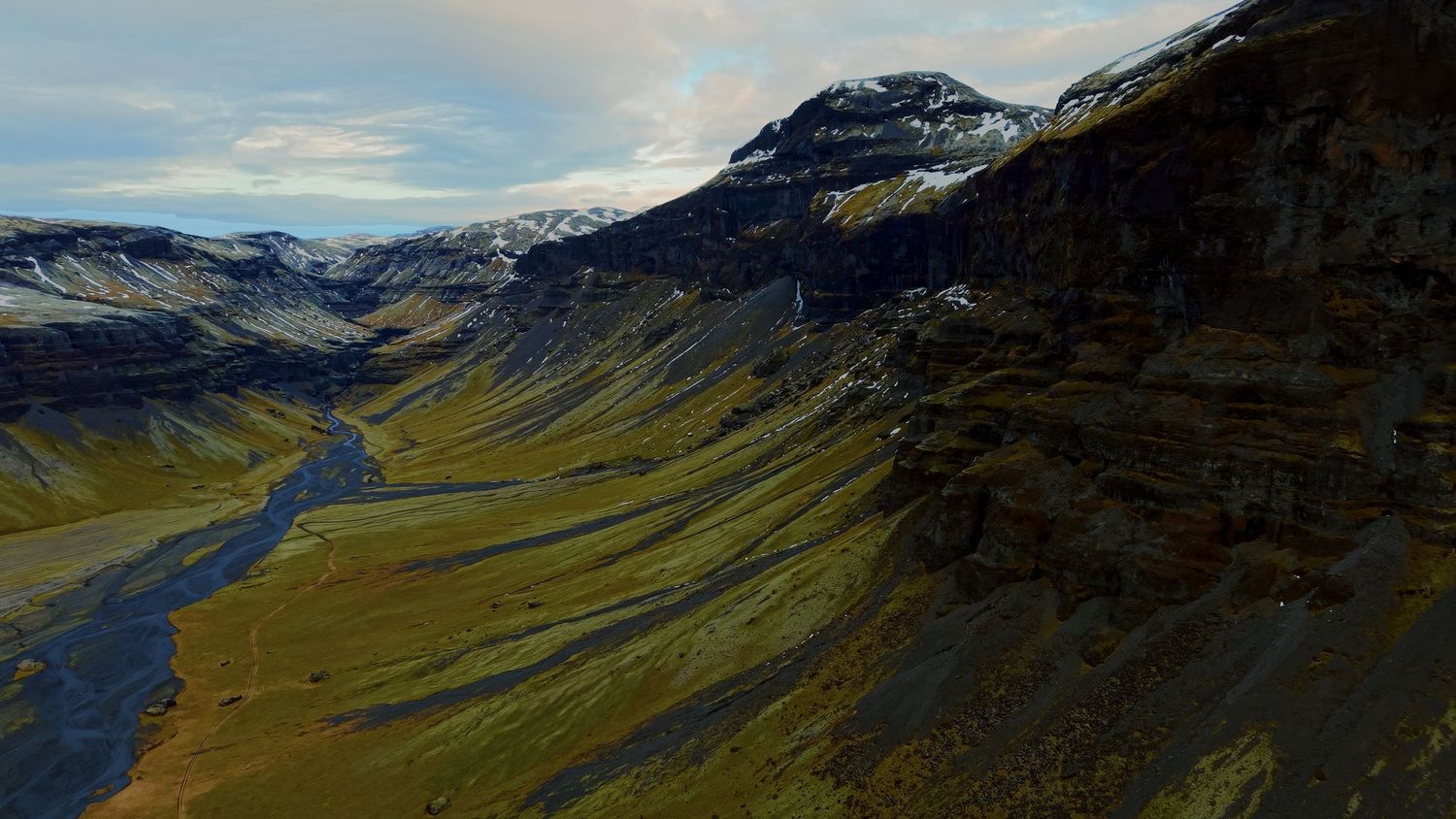 Panoramic aerial view of a winding river valley surrounded by rugged mountain peaks and green slopes in a remote landscape.