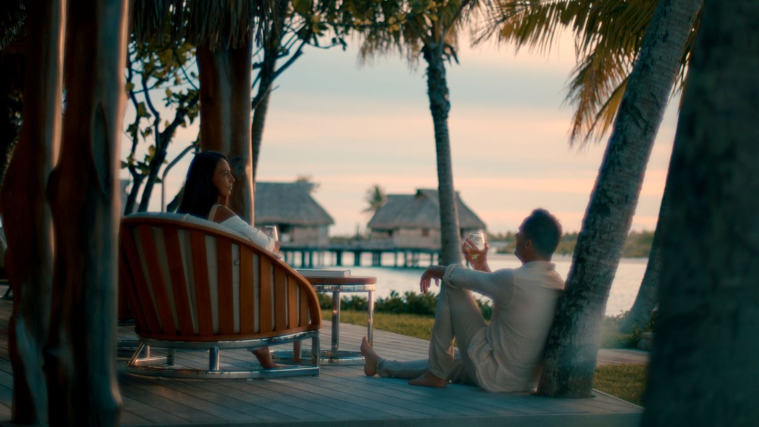 Wooden beach chairs and umbrellas line a tropical shoreline at sunset with palm trees swaying in the breeze.