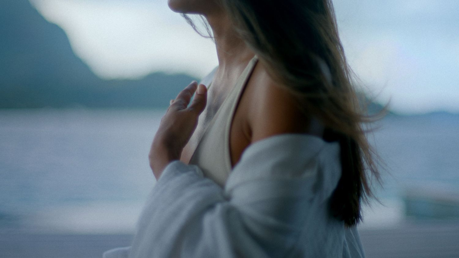 Woman in white robe standing by ocean with mountain backdrop, hair blowing in wind.