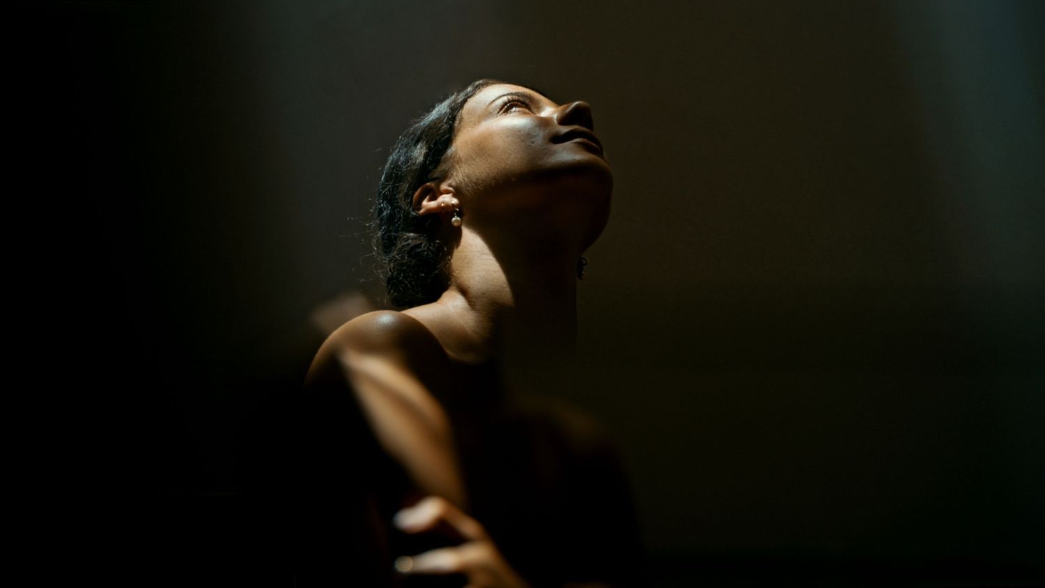 A dramatic close-up portrait in dark lighting showing a person with closed eyes tilting their head upward.