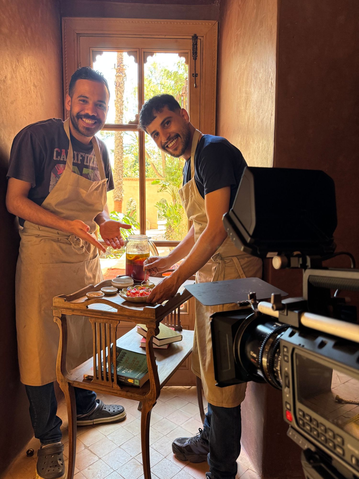 Two people wearing aprons prepare food on a wooden table near a window while being filmed with a professional camera setup.
