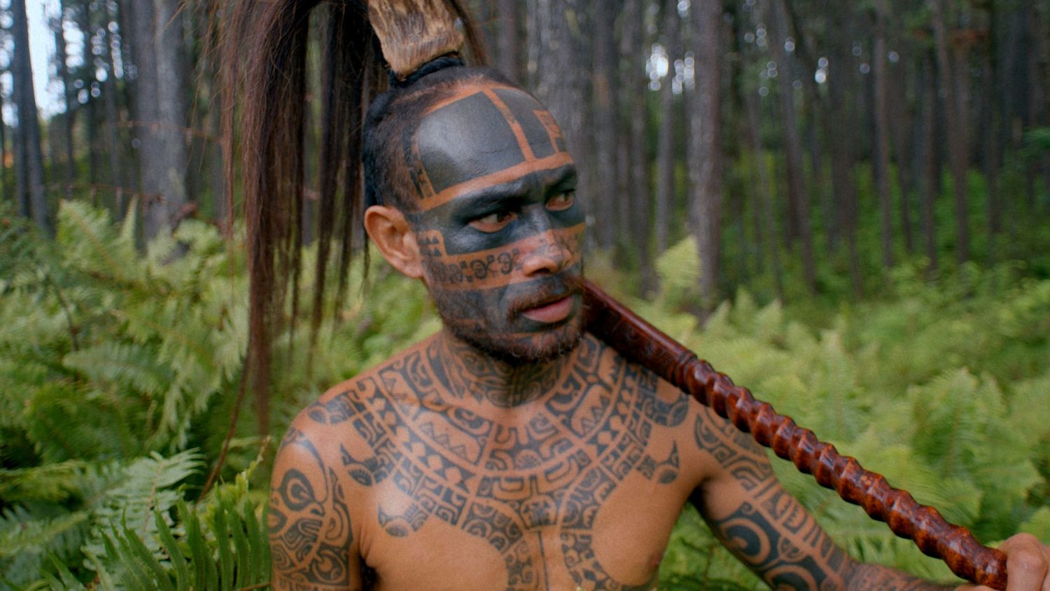 Maori warrior with traditional facial tattoos and tribal body art poses in a lush forest setting.