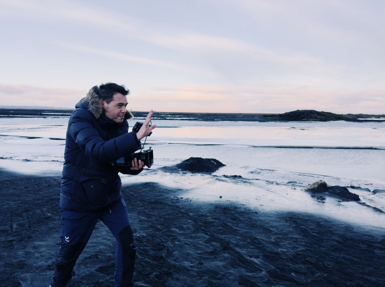 Photographer in a navy winter coat taking photos on a snowy beach at sunset.