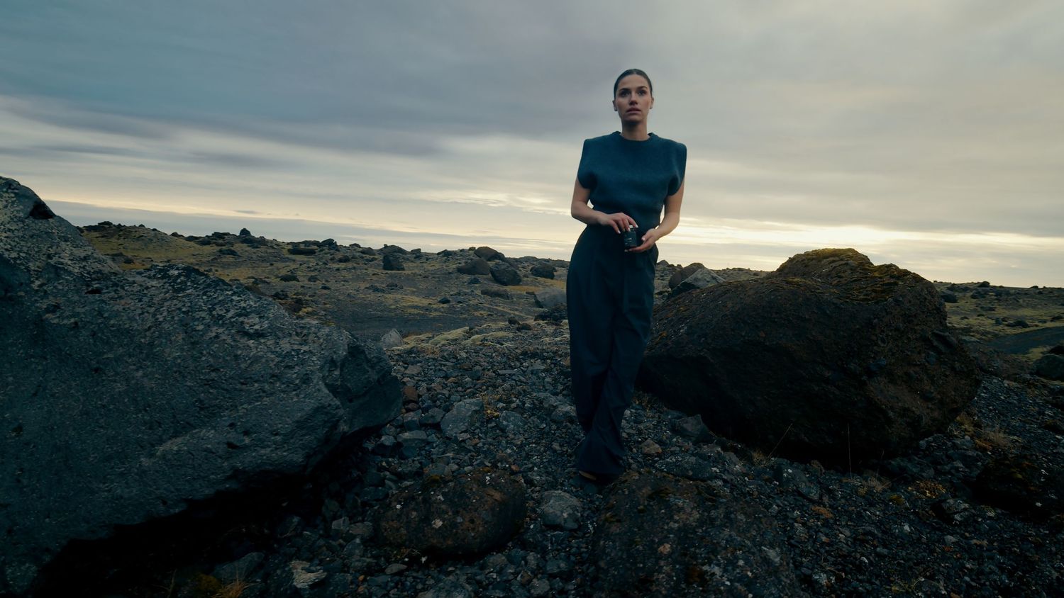 A person stands on rocky volcanic terrain overlooking a dramatic cloudy landscape at sunset.