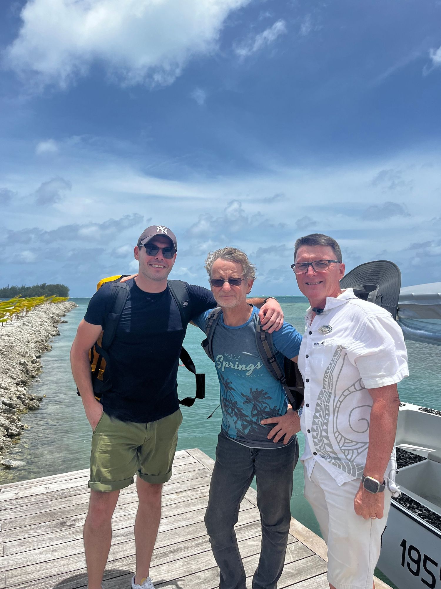 Three people standing together on a wooden dock by the ocean on a sunny day with blue skies and boats visible.