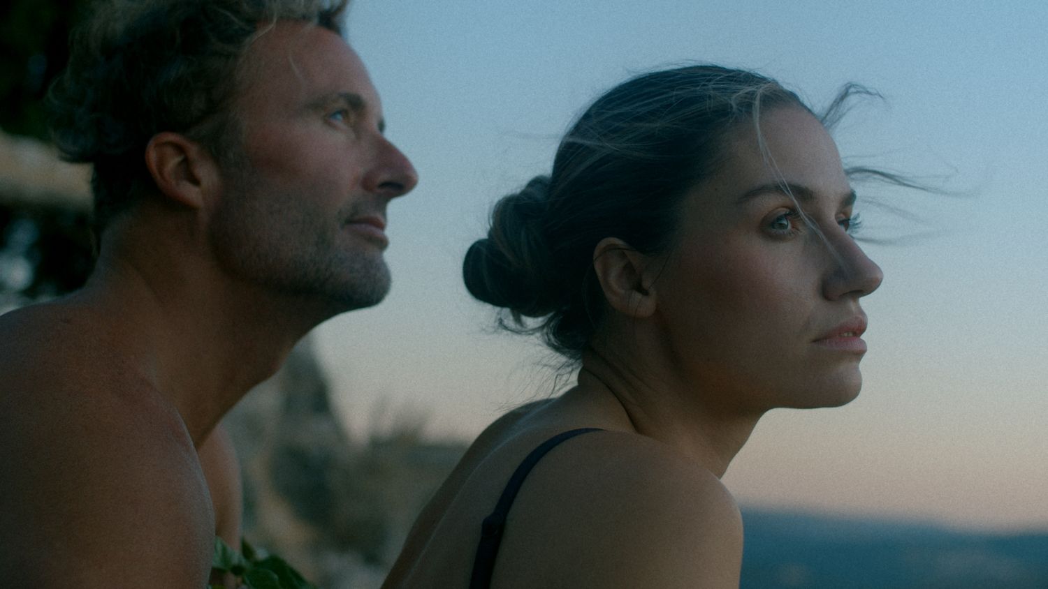 A man and woman stand together outdoors at dusk, gazing into the distance with a scenic hillside backdrop.