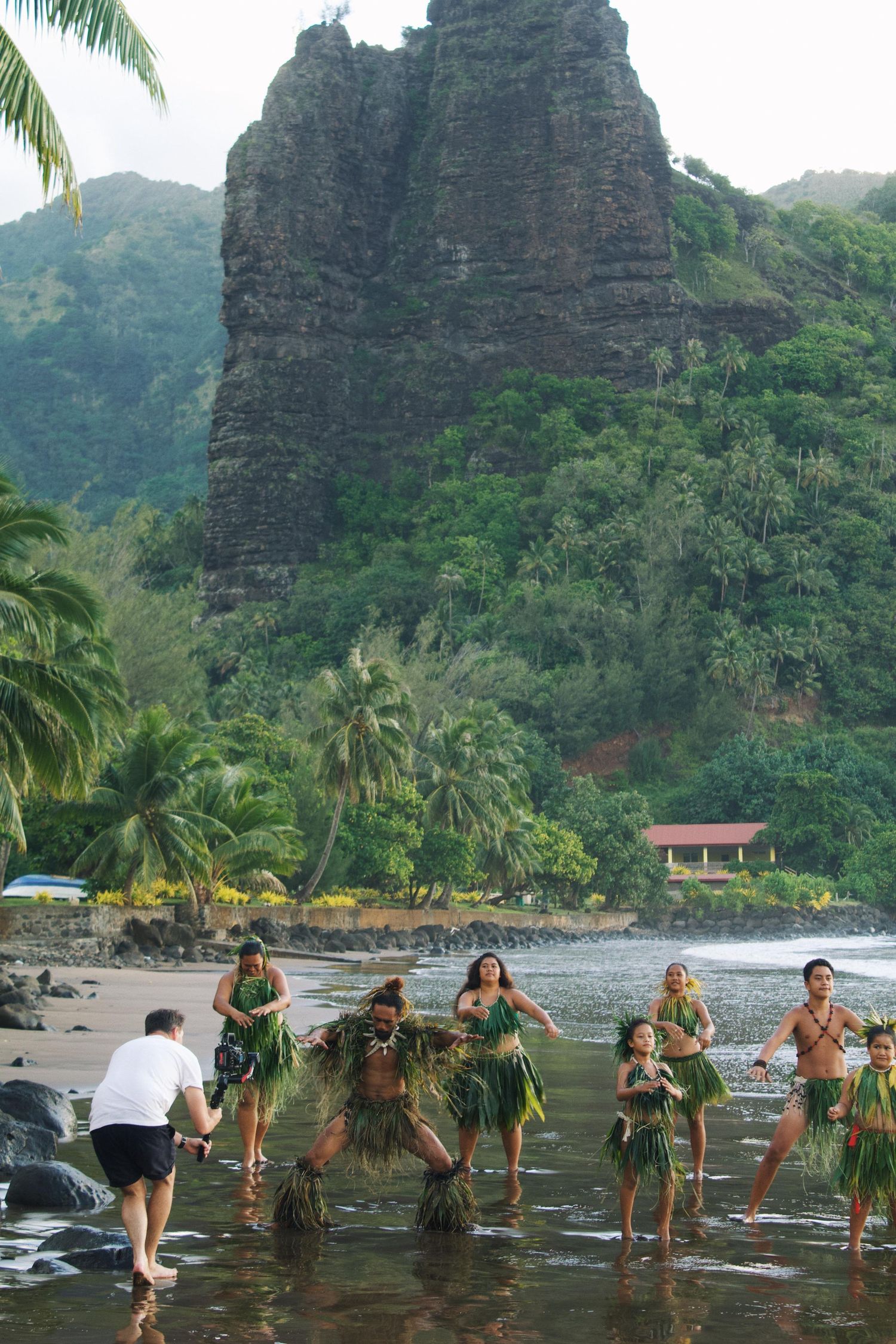 Traditional Polynesian dancers in grass skirts perform on a tropical beach with lush green mountains in the background.