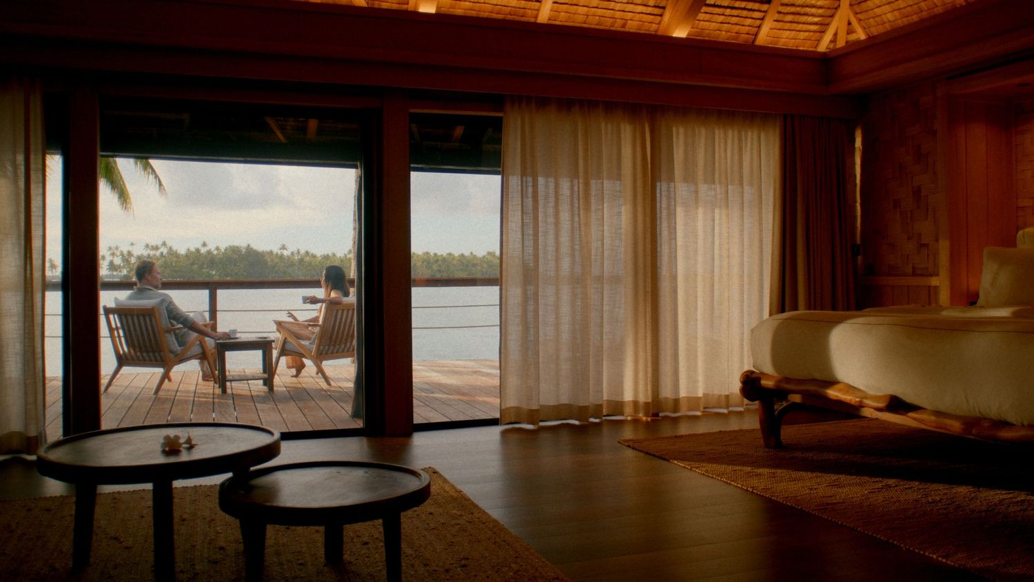 Luxurious overwater bungalow interior with sheer curtains, teak floors, and panoramic views of a tropical lagoon.