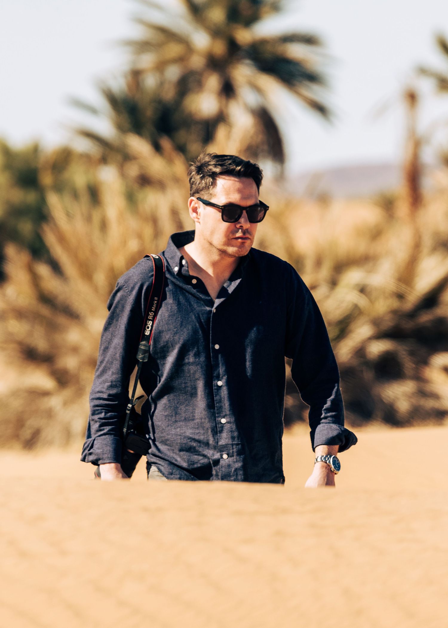 A person in sunglasses and dark button-down shirt walks through a desert landscape with palm trees in the background.