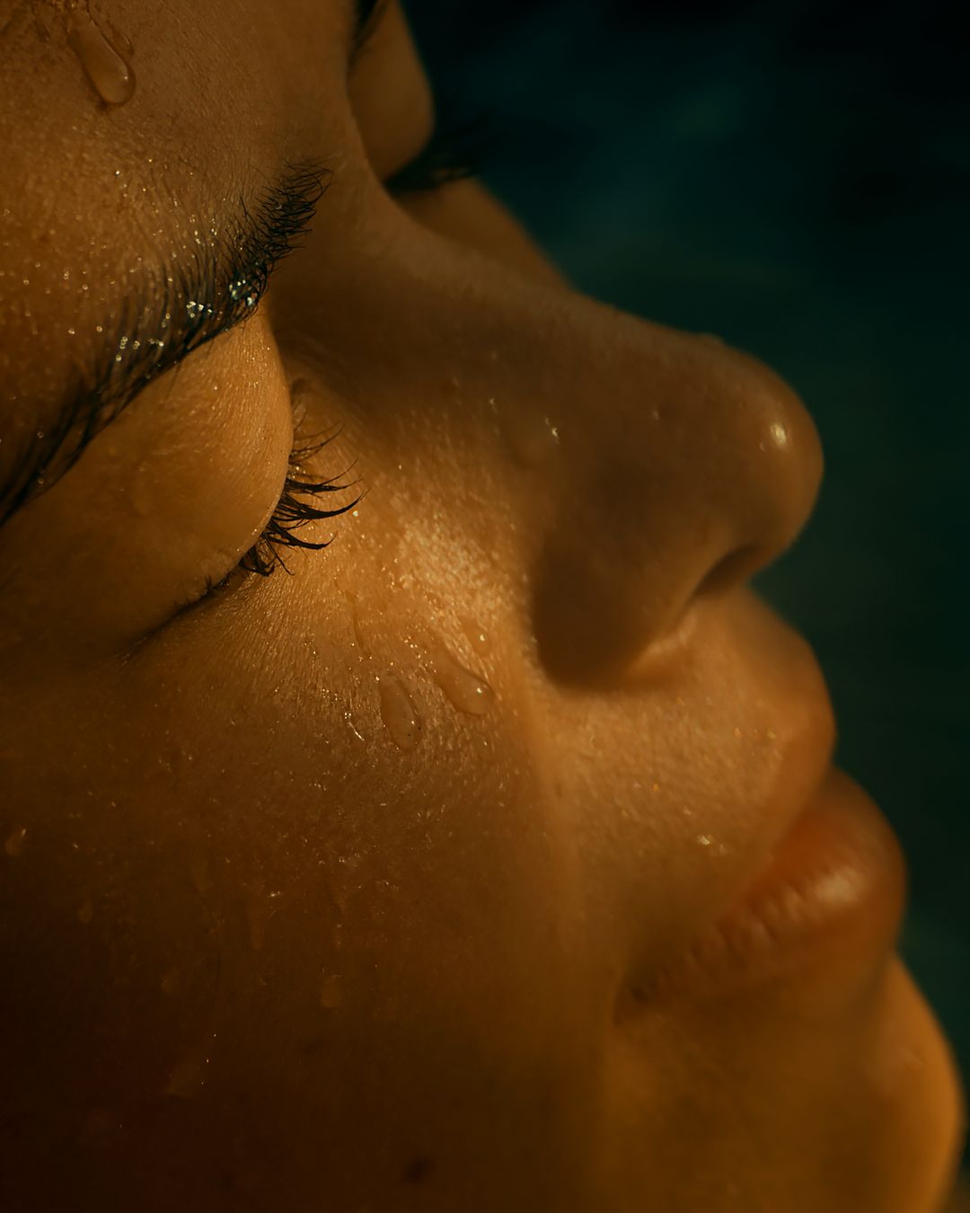 Close-up profile view of a dewy, glowing face with long eyelashes against a dark background.