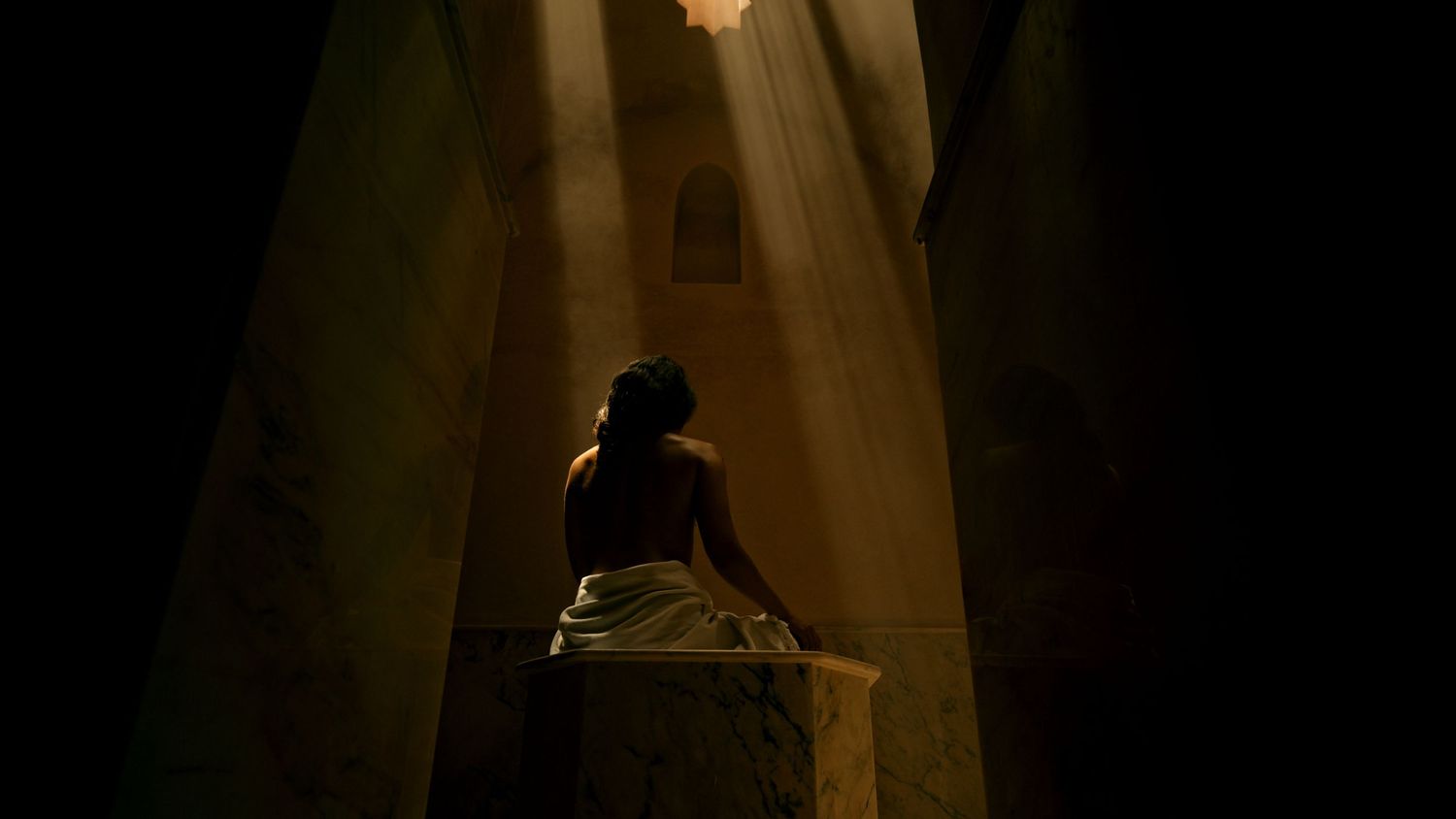 A dark silhouetted figure stands in dramatic light beams shining down through a misty or smoky atmosphere.