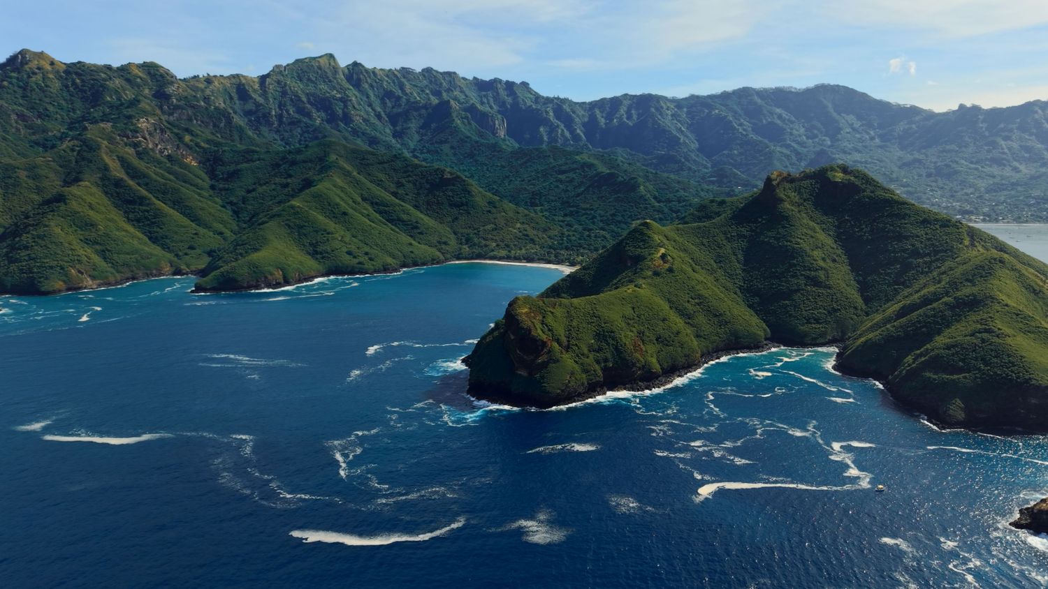 Aerial view of lush green mountainous coastline with turquoise waters and ocean waves in a tropical island setting.