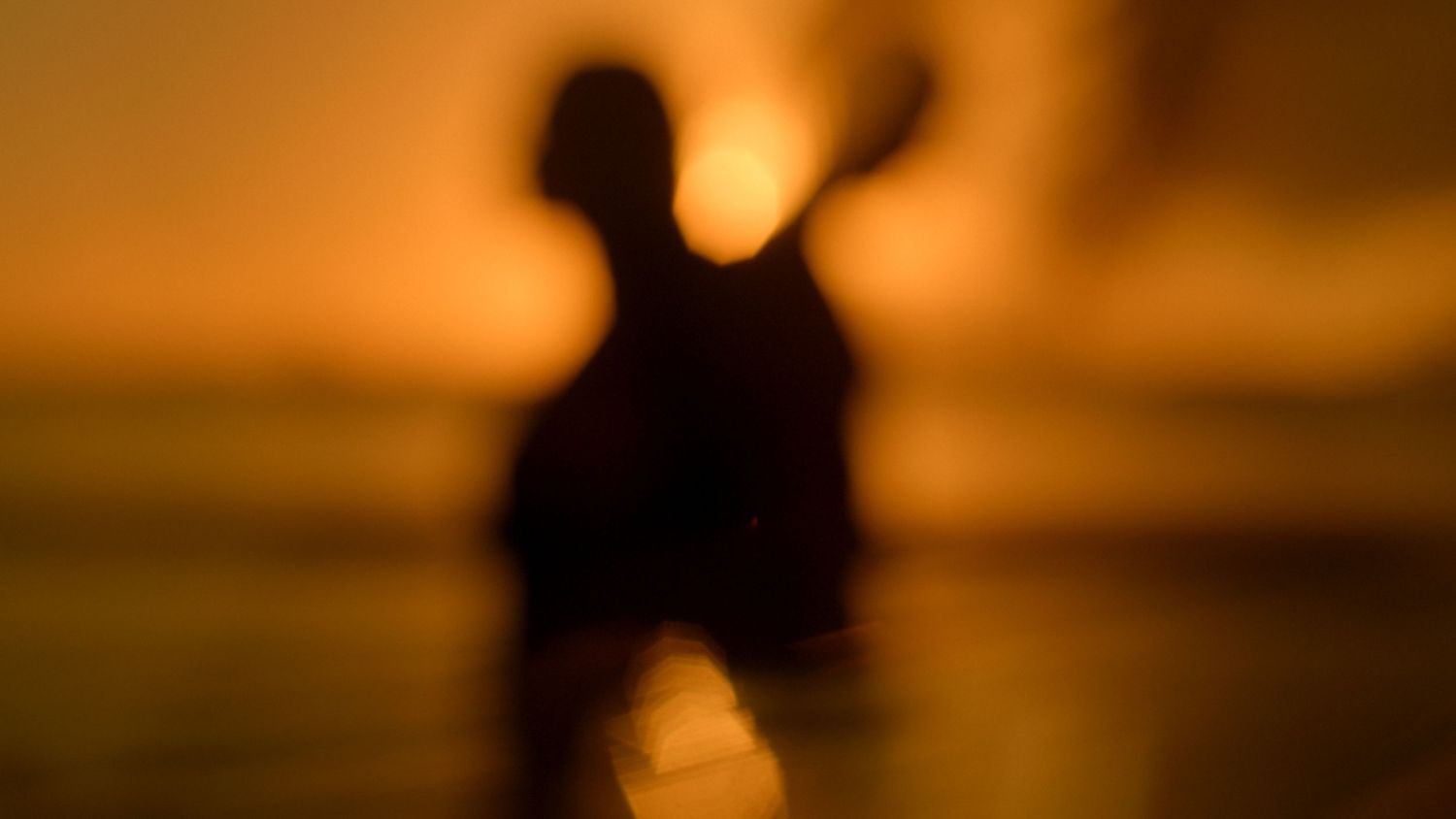 Blurry silhouette of a person against a warm golden sunset reflecting on calm water.