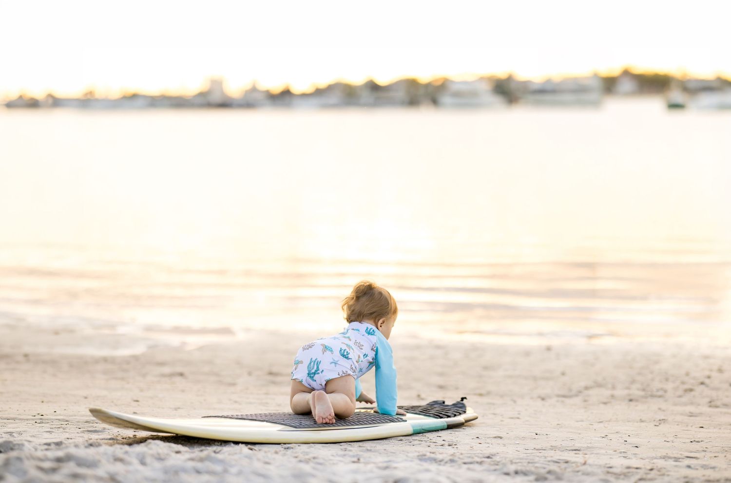 A baby in a floral rashguard crawls on a surfboard on a sandy beach at sunset.