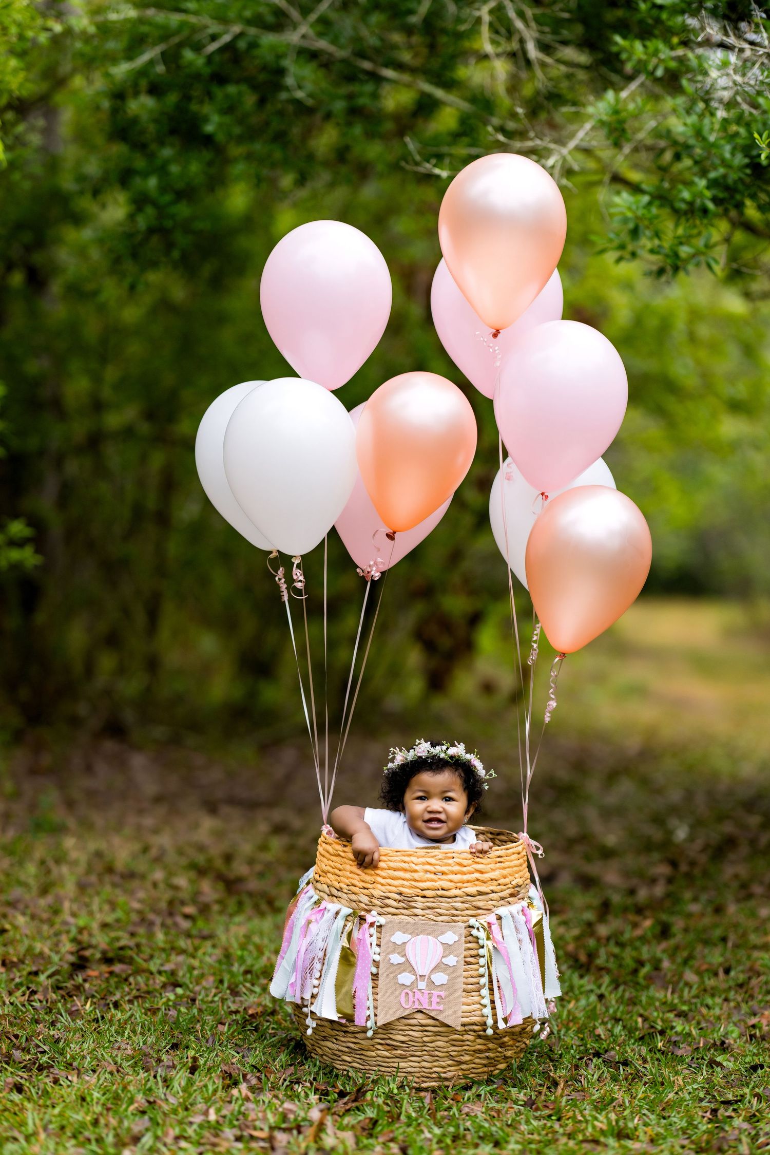 Creative first birthday photo with pink and peach balloons attached to decorated basket outdoors.