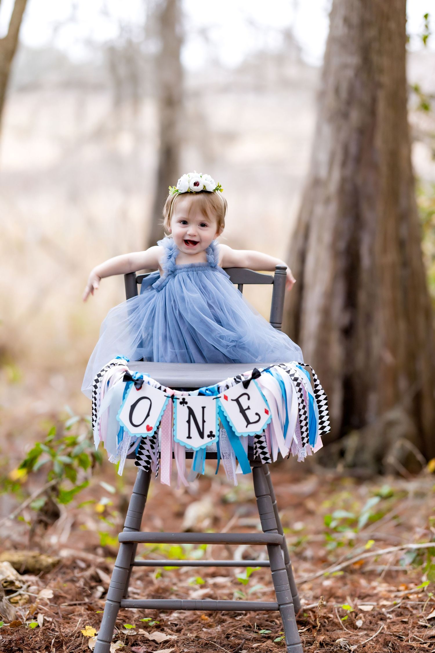 First birthday photo with toddler in blue tulle dress on grey chair with decorative ONE banner.