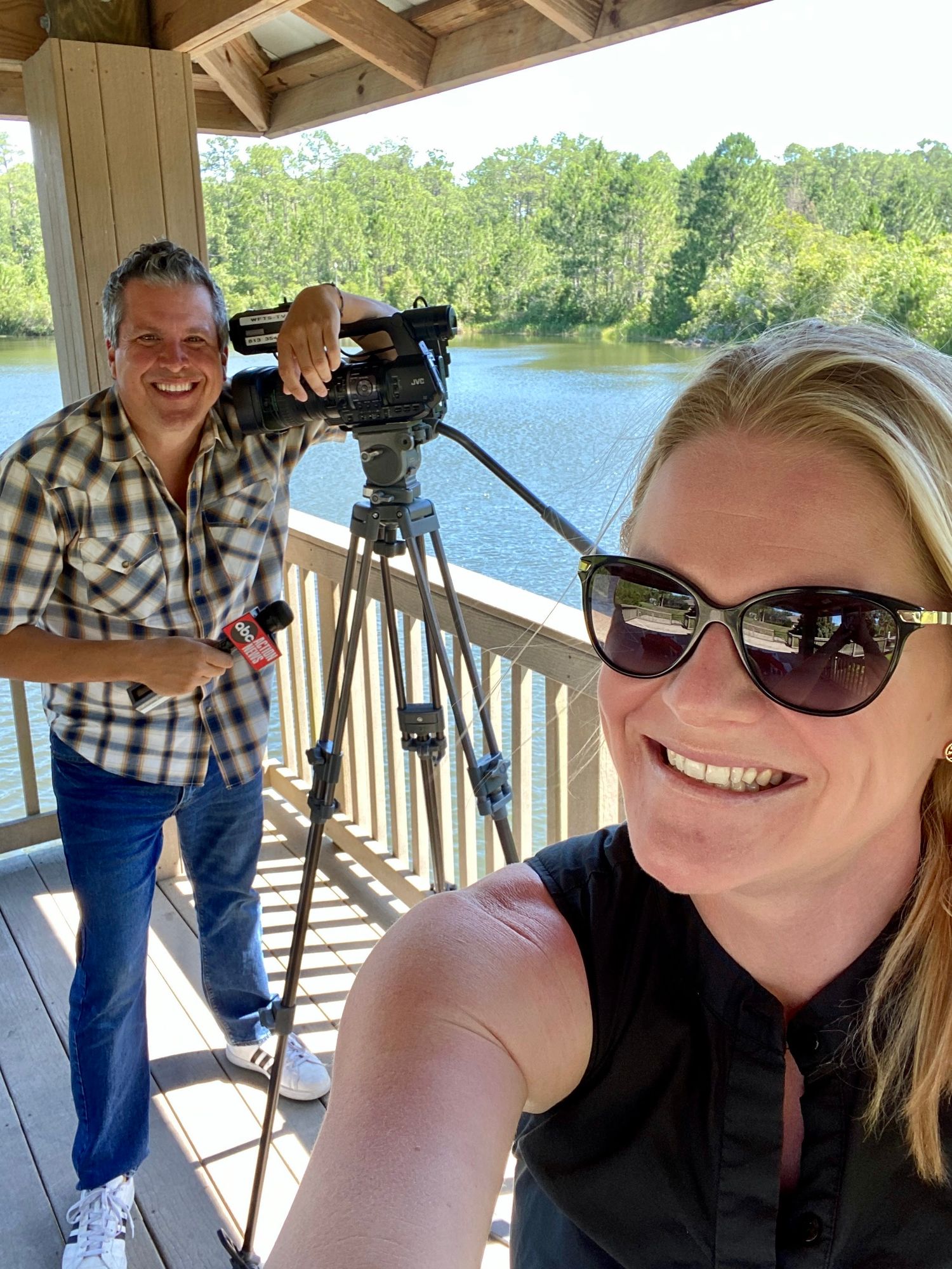 Two people taking photos from a covered deck overlooking a scenic lake with trees in the background.