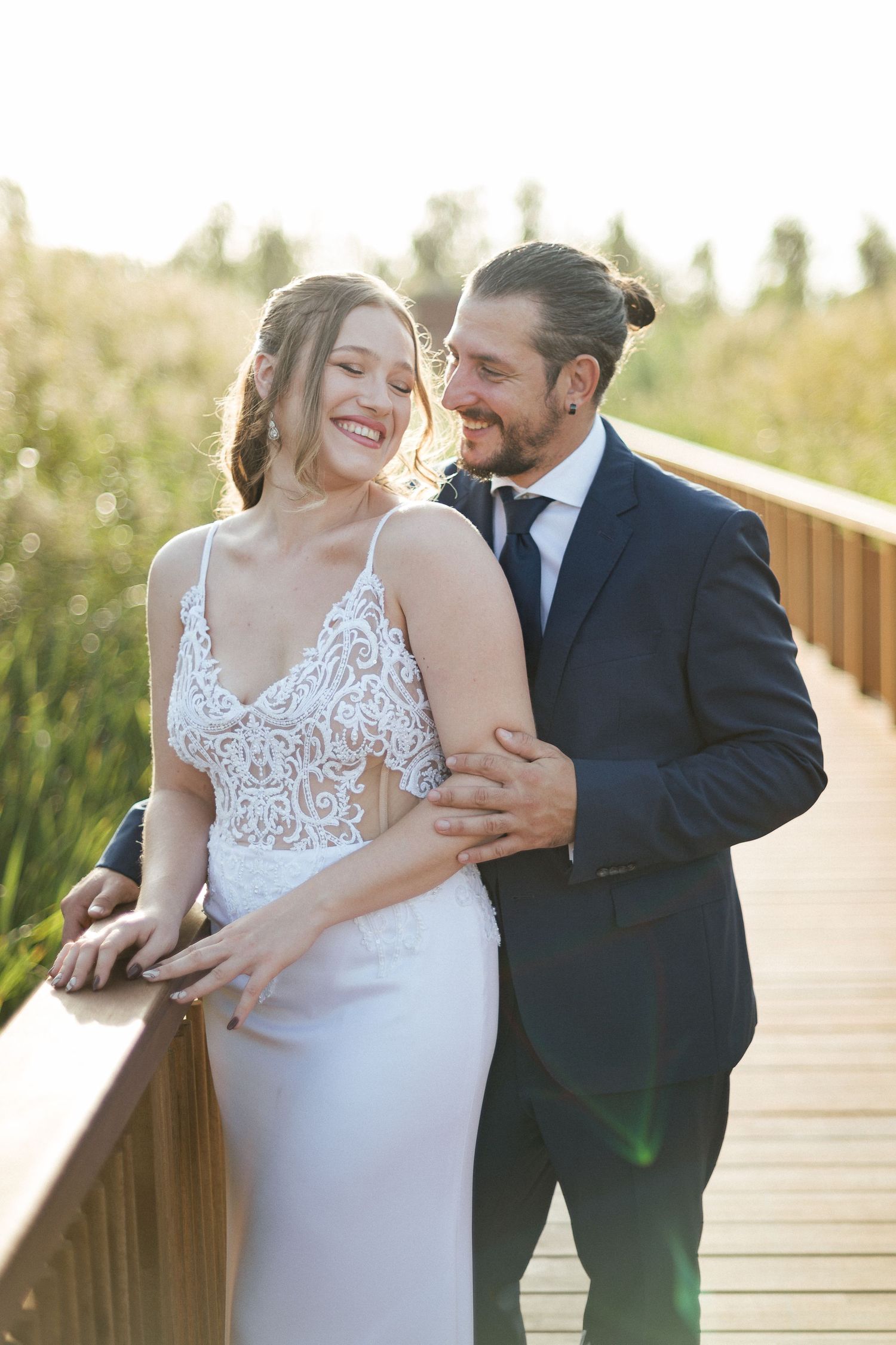 Joyful wedding couple sharing a tender moment on a wooden boardwalk surrounded by summer grass.