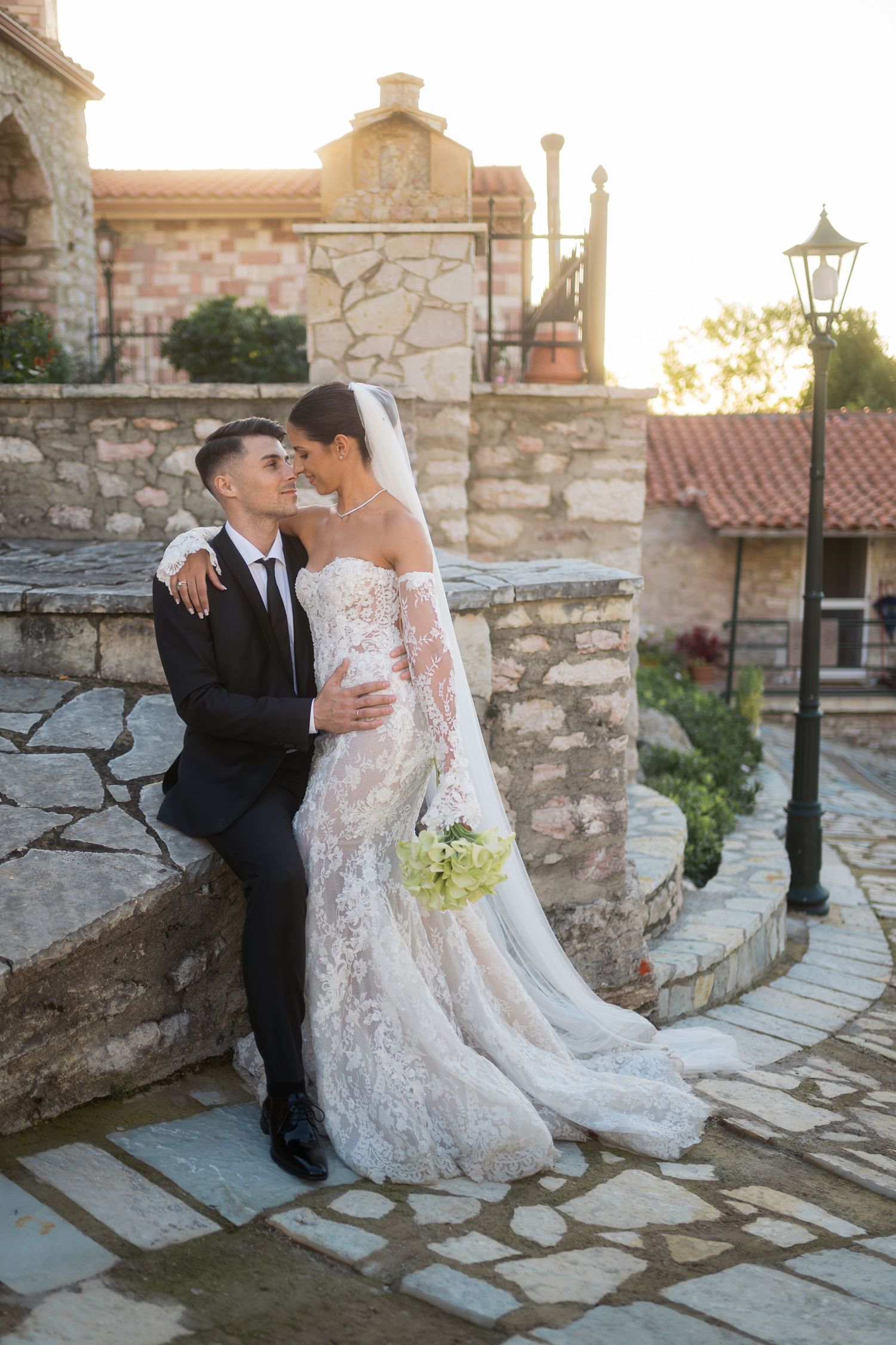 Newlywed couple shares romantic moments on stone steps at sunset in a Mediterranean villa setting.