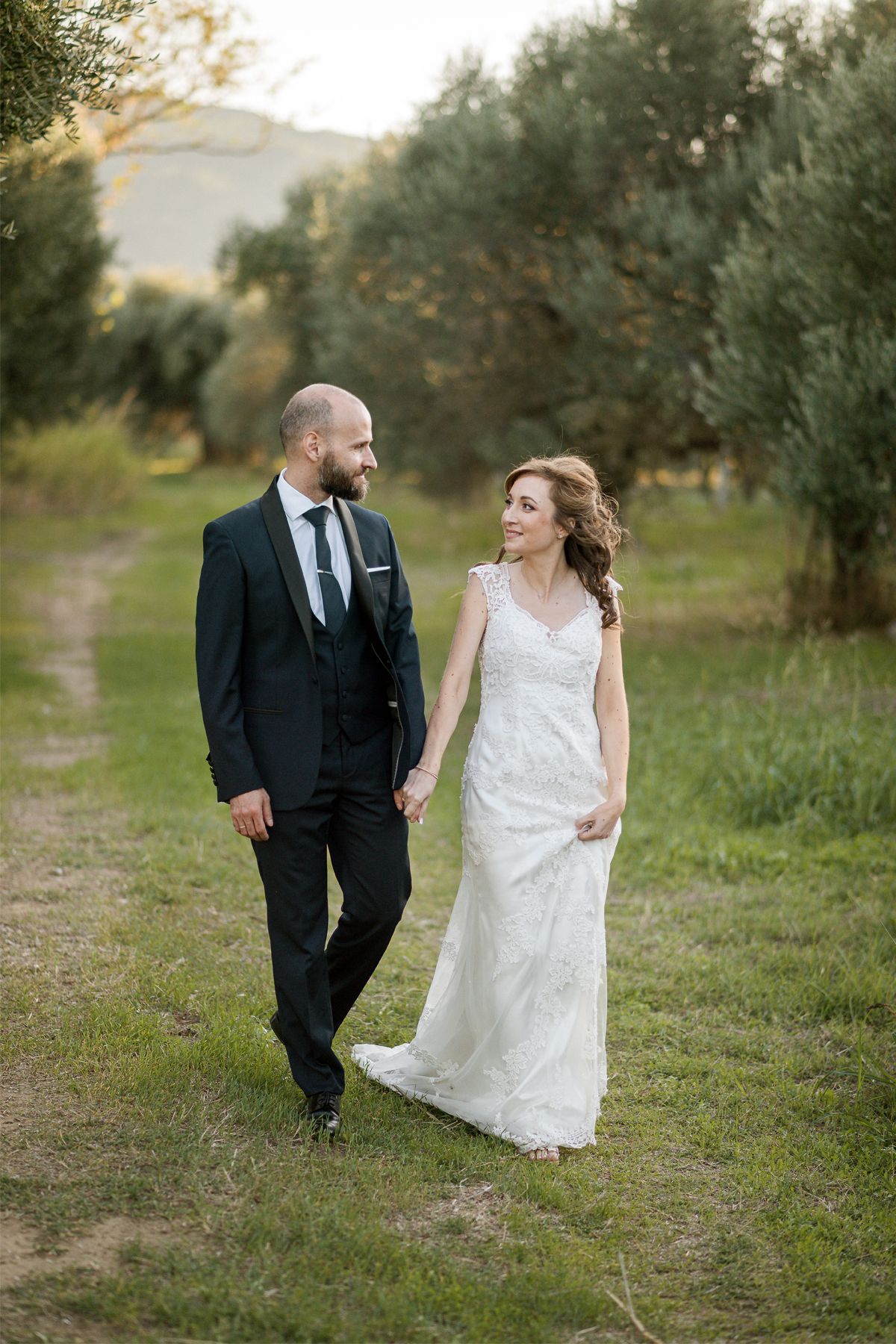 A bride in a lace wedding dress and groom in dark suit walking together on a path surrounded by olive trees.
