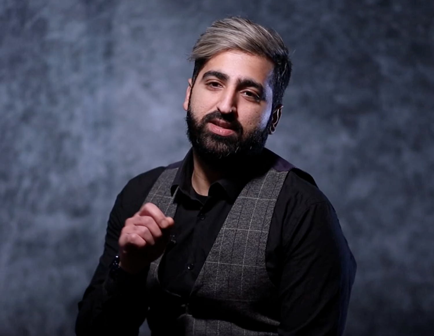 A bearded man in a black shirt and plaid vest poses confidently against a textured gray backdrop.
